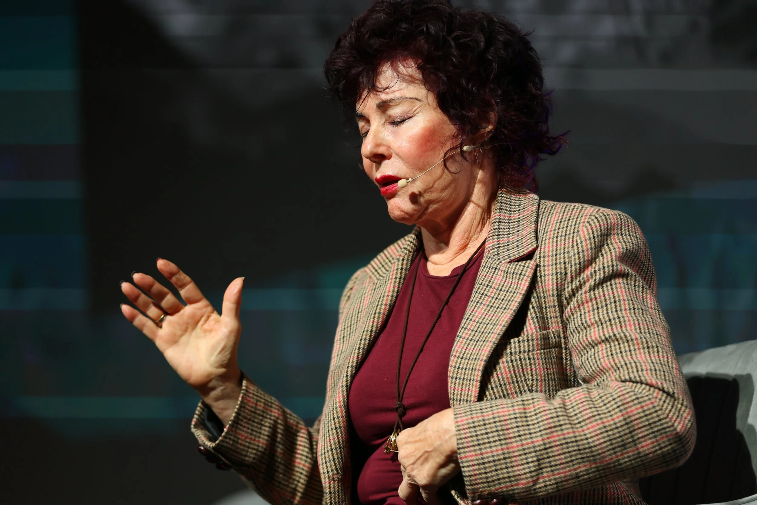 A woman with dark, curly hair wearing a plaid blazer and maroon shirt, sitting with her eyes closed and speaking into a headset microphone, gesturing with her right hand.