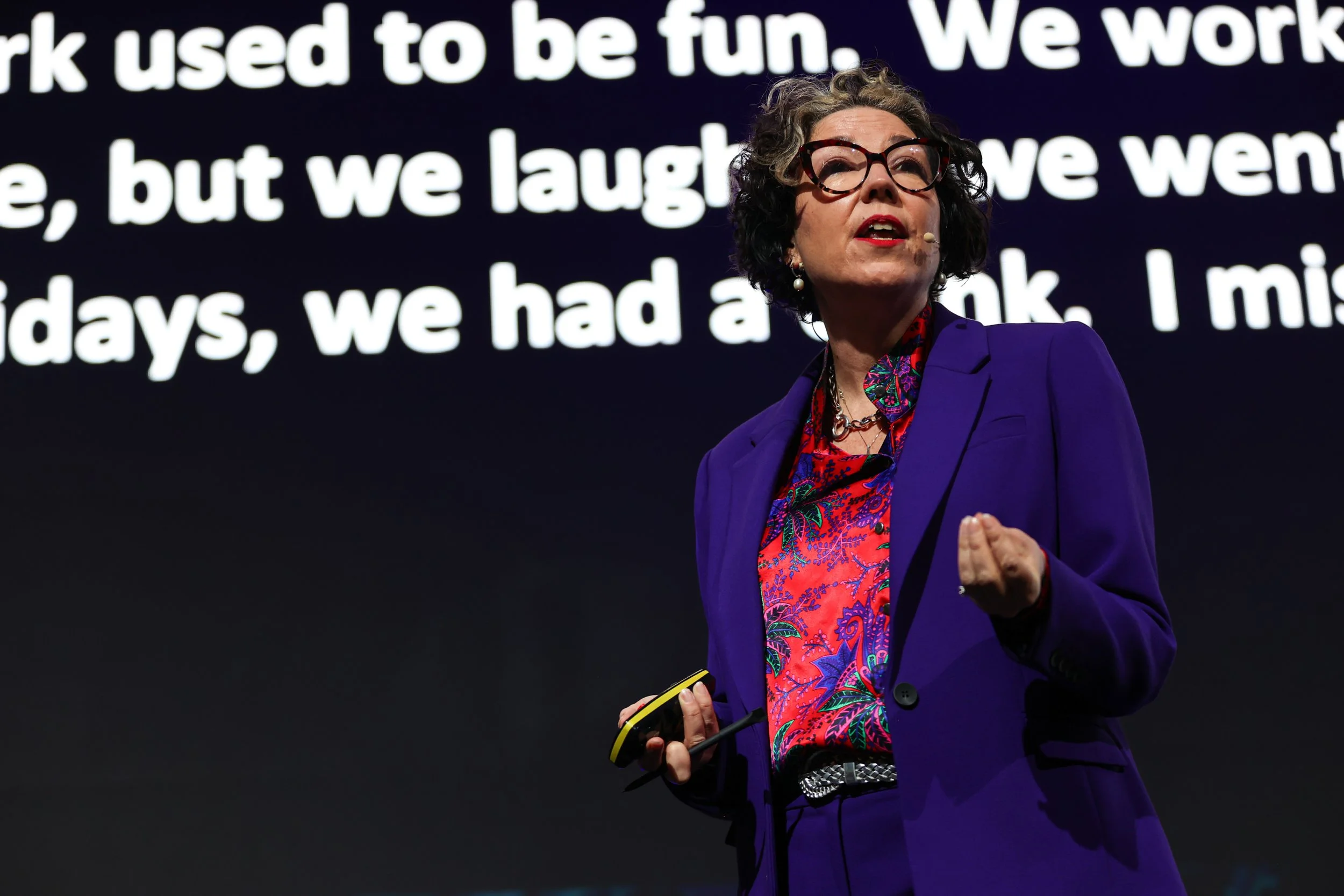 A woman with glasses, curly hair, and earrings giving a presentation or speech on stage, wearing a purple blazer over a colorful patterned shirt, holding a yellow smartphone, with a large screen displaying text behind her.