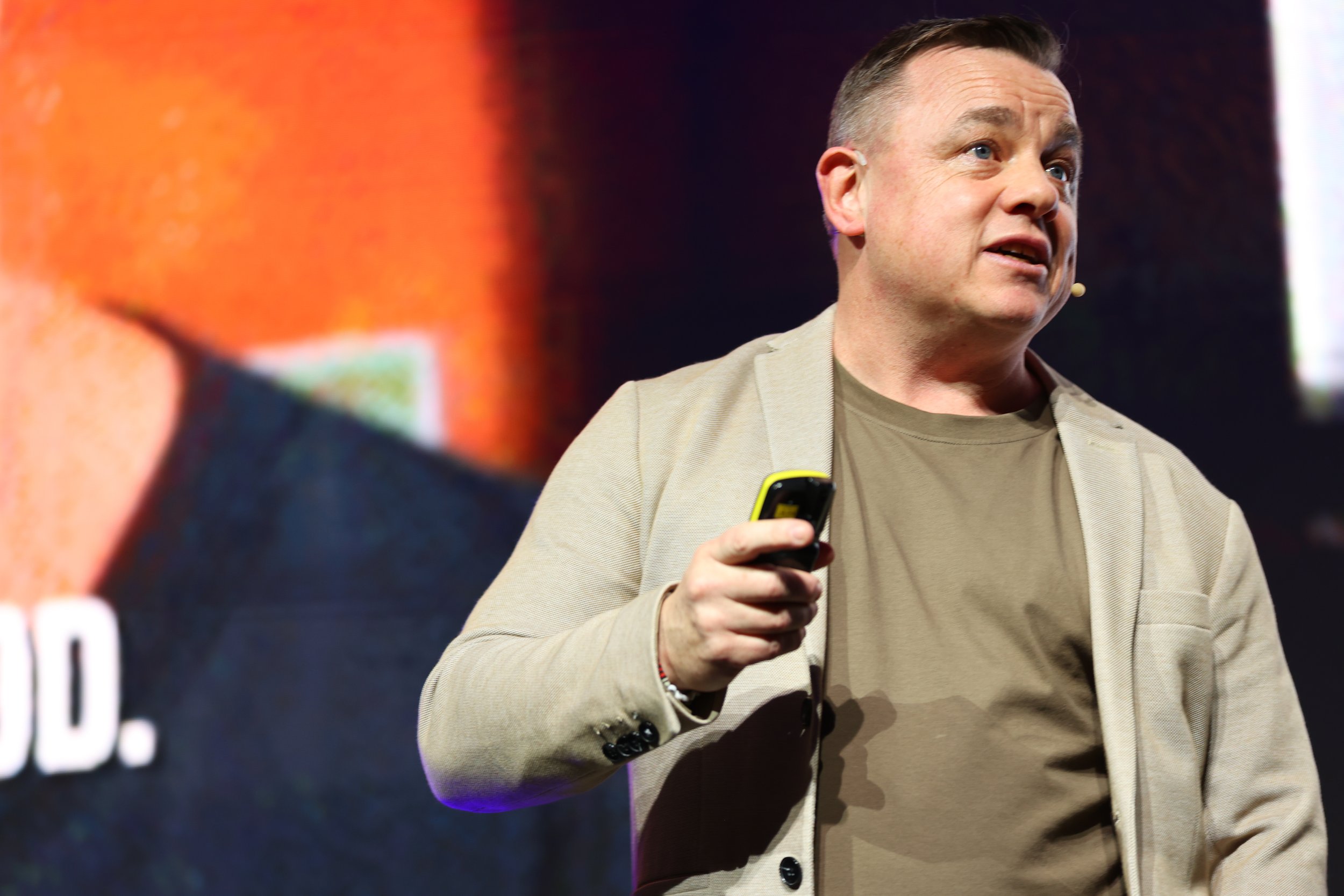 A man in a beige jacket and brown shirt speaking on stage, holding a yellow device, with a dark background and a large screen behind him.