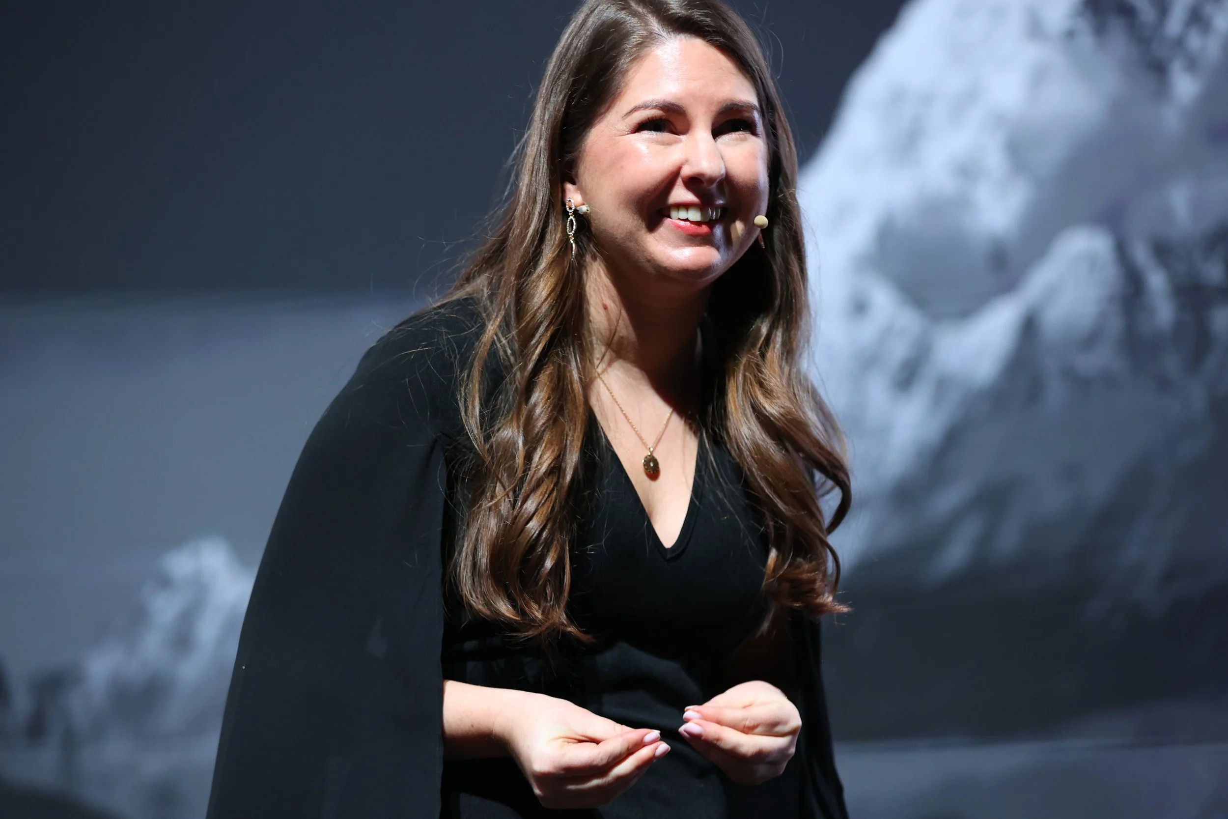 A woman with long wavy brown hair, wearing a black dress and jewelry, smiling on stage with a mountain landscape backdrop.