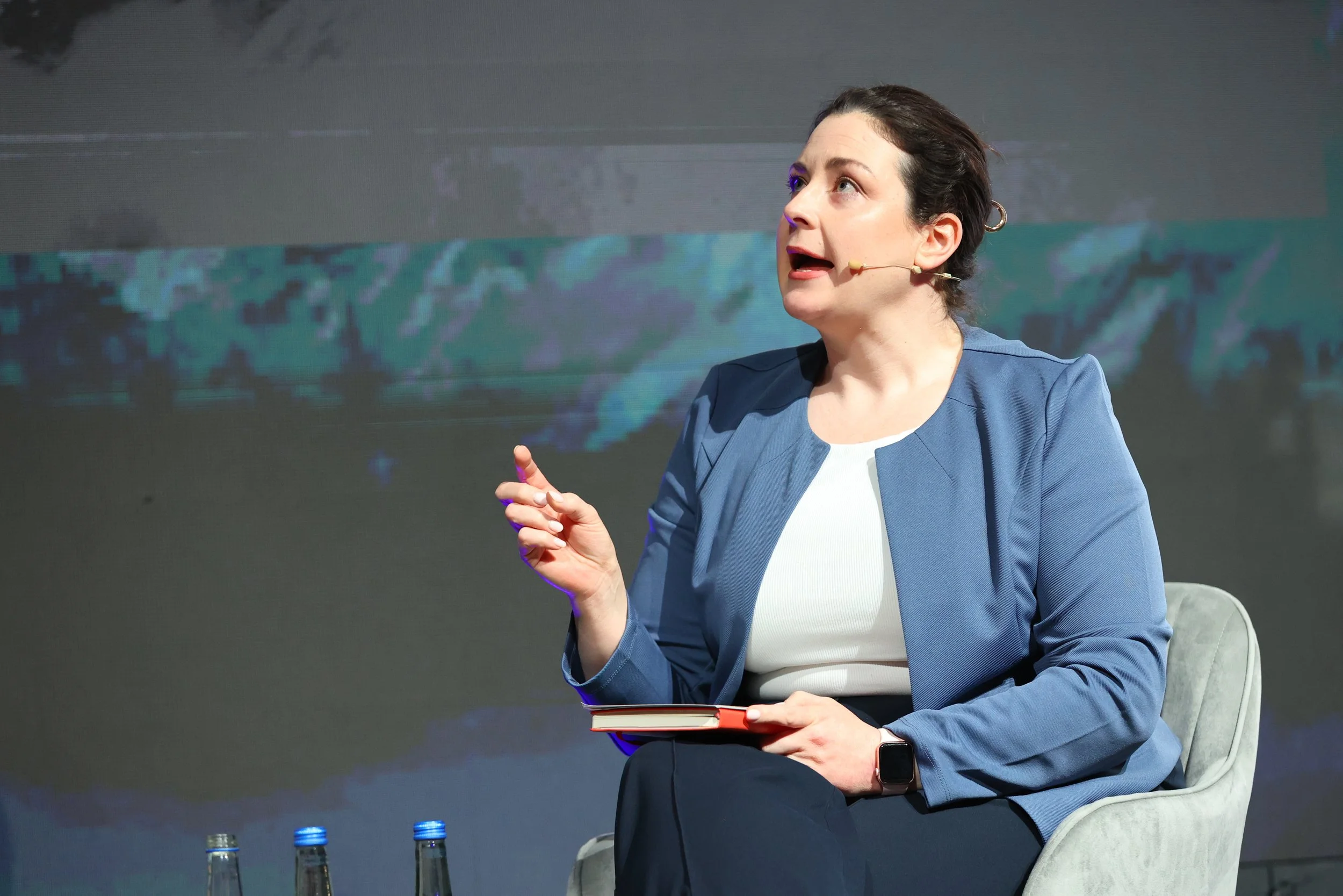 A woman with dark hair in a bun wearing a blue blazer and white top, speaking into a headset microphone while seated on a gray chair, holding a notebook, with a dark patterned background.