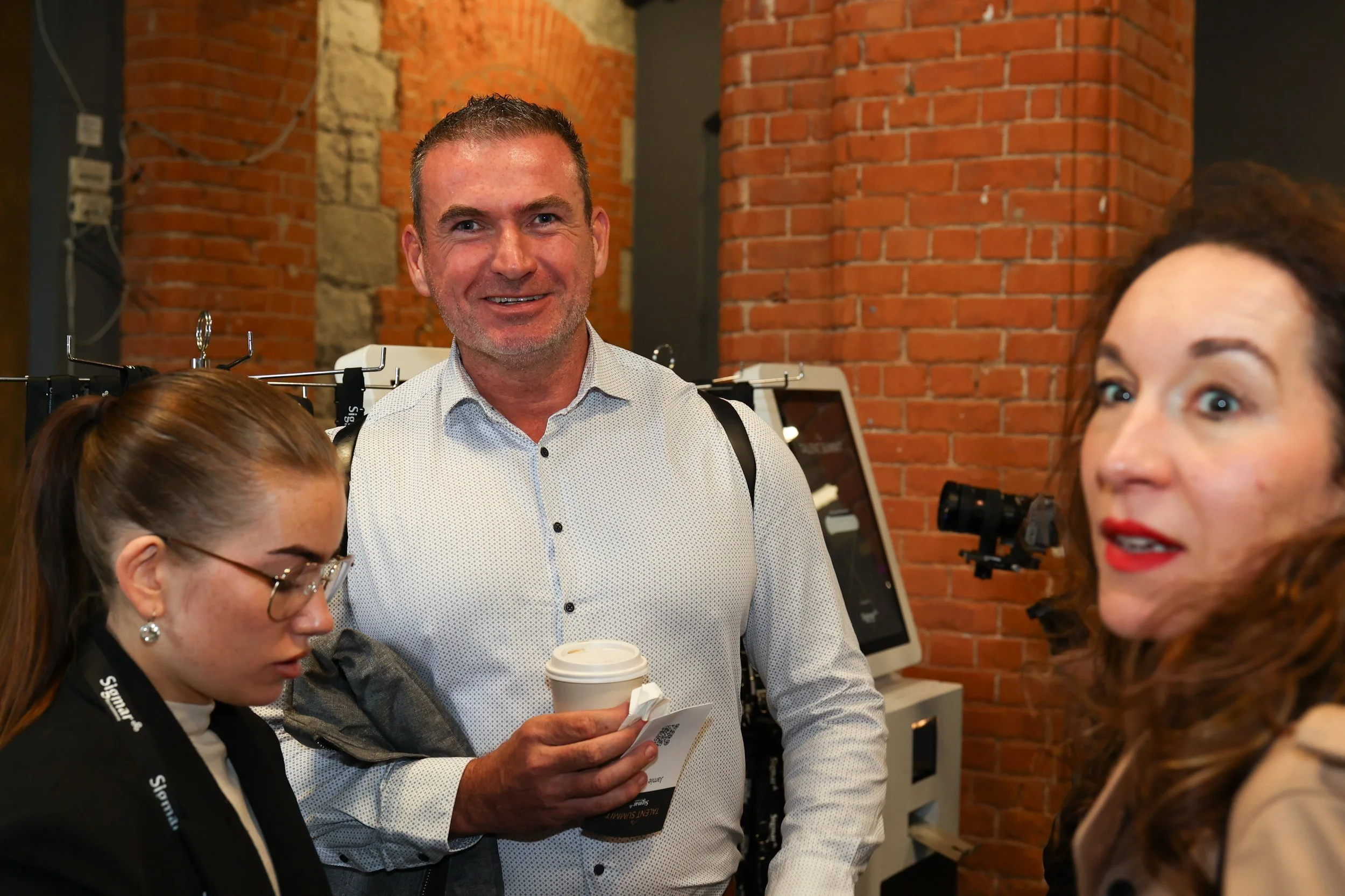 Three people at an indoor event with brick walls, a woman with glasses and a black lanyard looking down, a man smiling with a coffee cup, and a woman with long dark hair speaking.