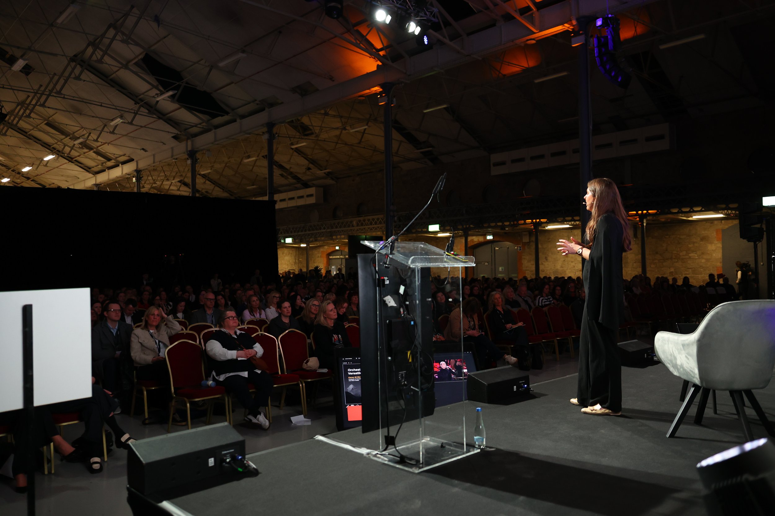 A woman in black clothing giving a presentation on stage in front of a large audience seated in a conference hall with high ceilings and exposed beams, illuminated by stage lights.