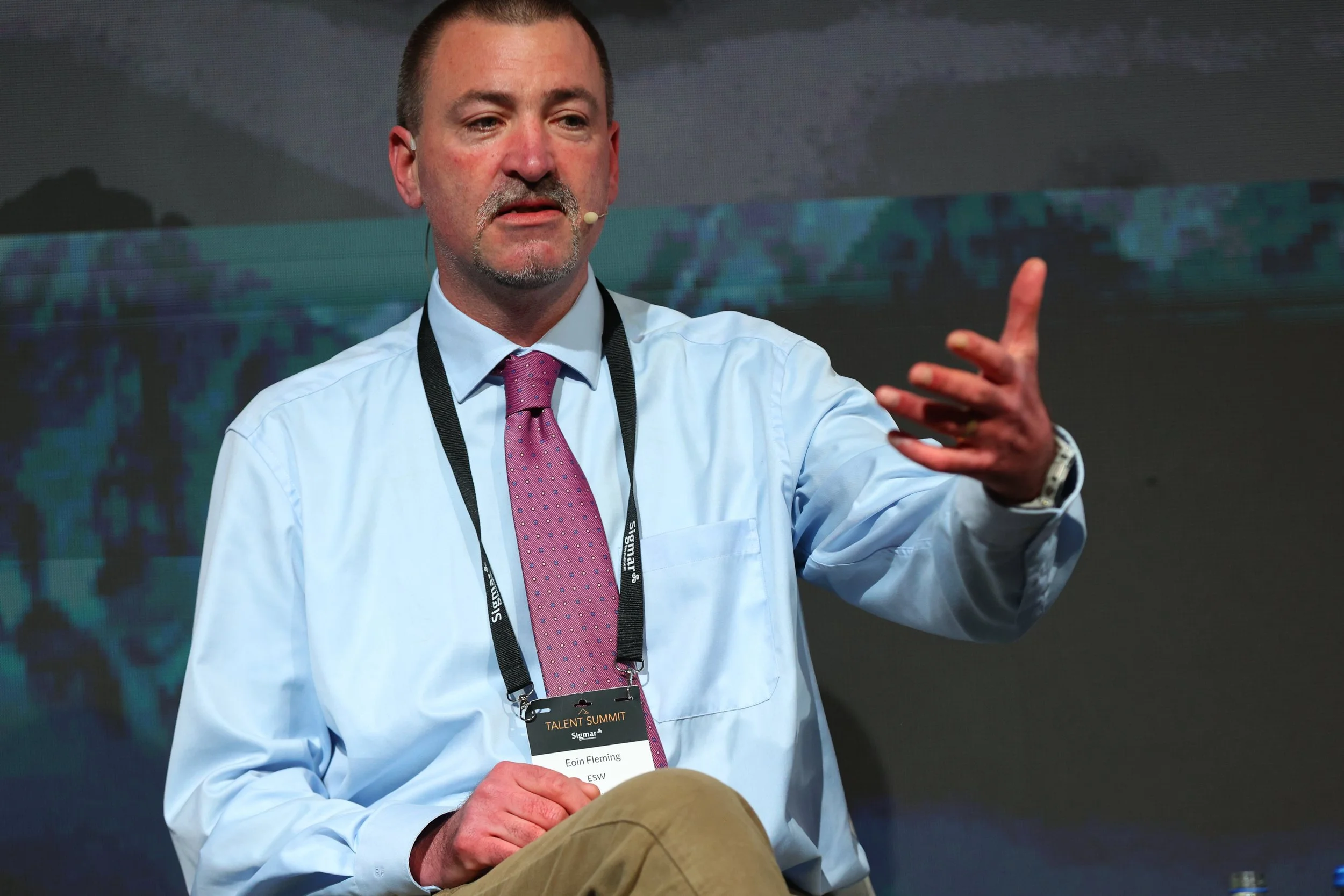 A man in a white shirt and pink tie, wearing a lanyard with a badge, is speaking on a stage. He is gesturing with his right hand.