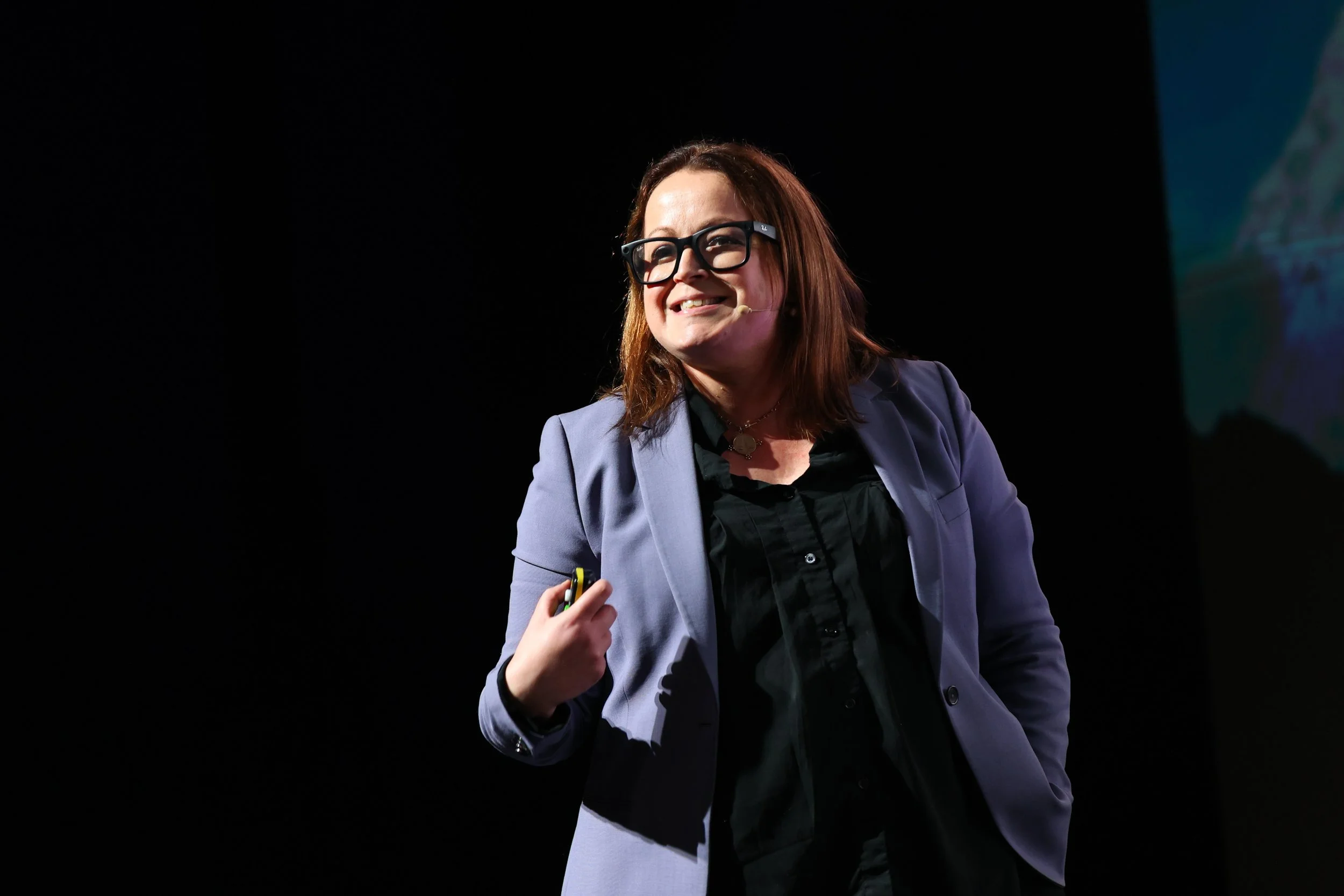 A woman with shoulder-length brown hair, wearing glasses and a lavender blazer, smiling while speaking on stage.