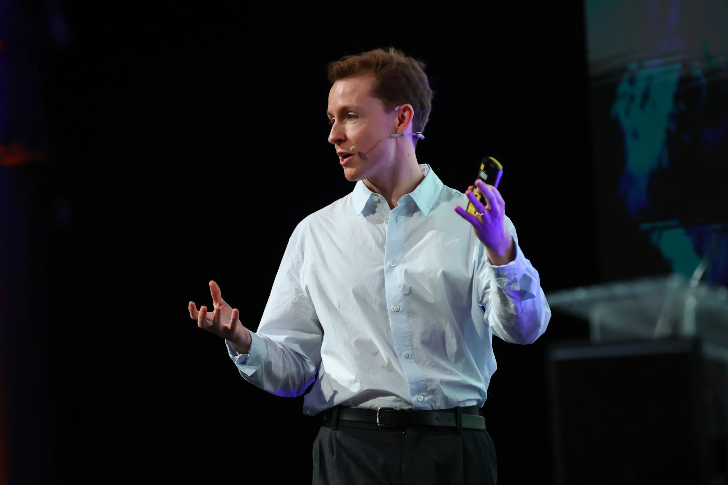 A man in a white shirt giving a presentation on stage with a dark background.