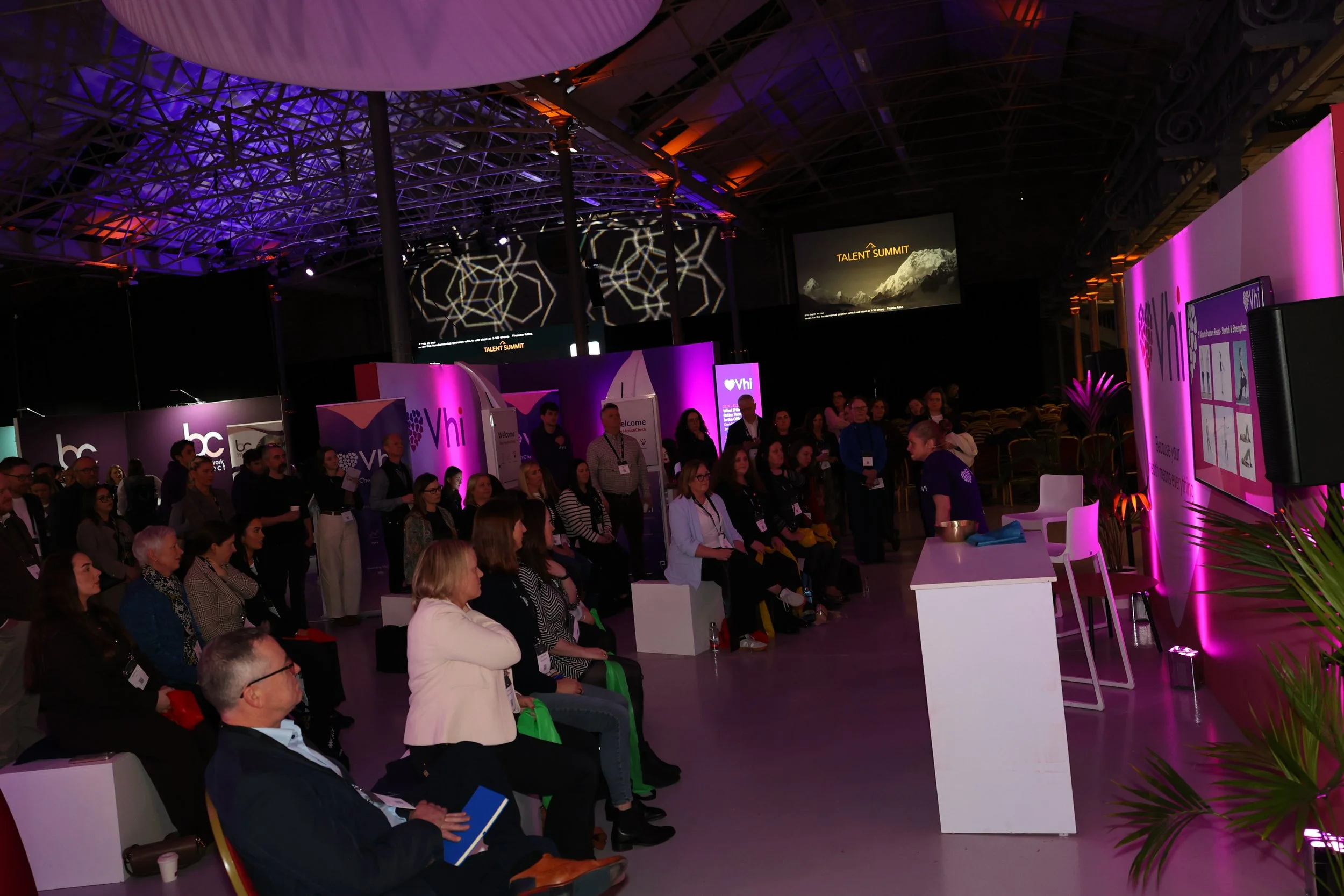 Audience attending a presentation at a conference in a large, dimly lit event space with purple lighting, stage screens displaying 'Talent Summit,' and a presenter speaking at a podium.
