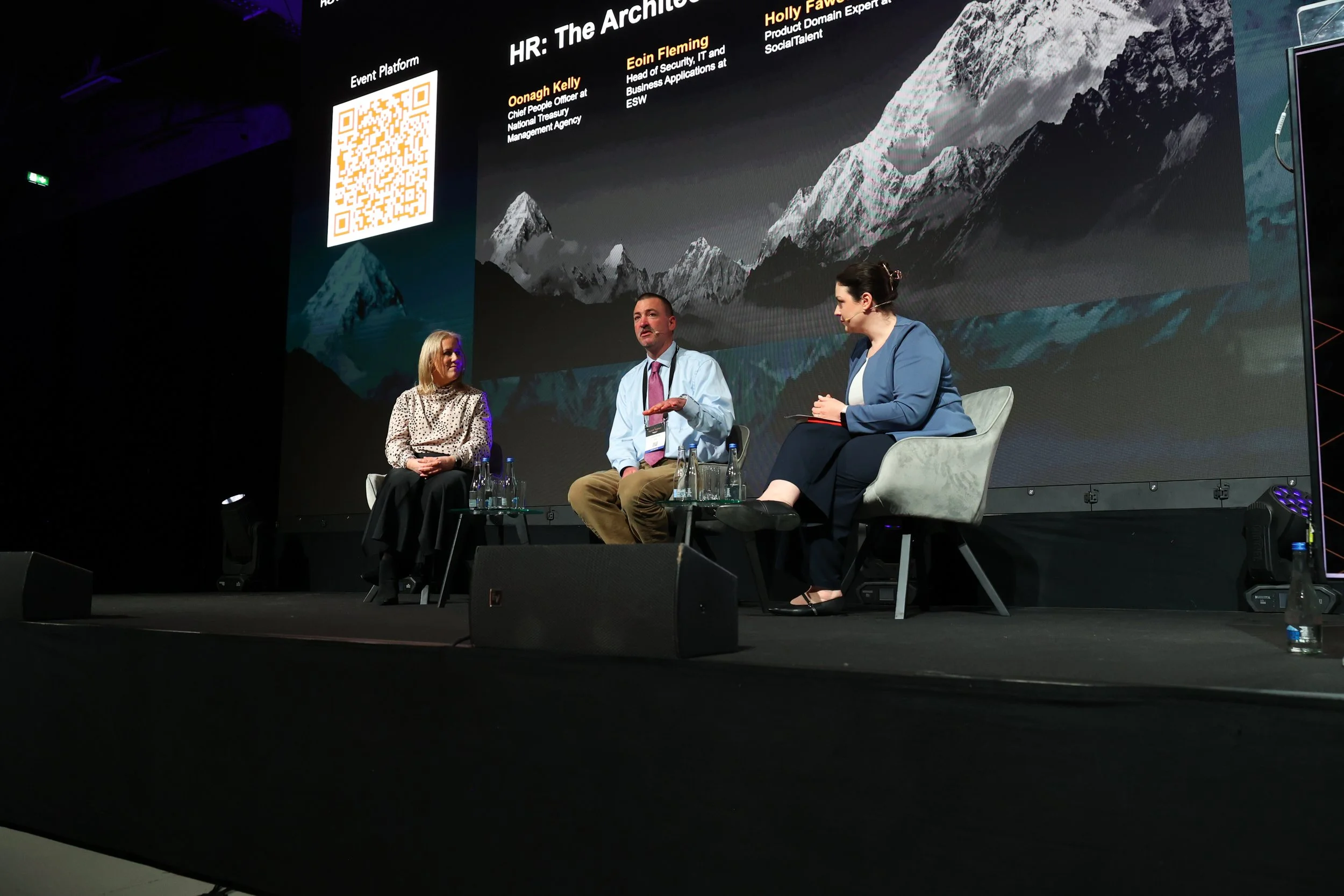 A panel discussion with three people seated on stage in front of a large screen displaying a mountain landscape and speaker information. The panel includes two women and one man, each with bottles of water on their tables. The man is speaking, and th