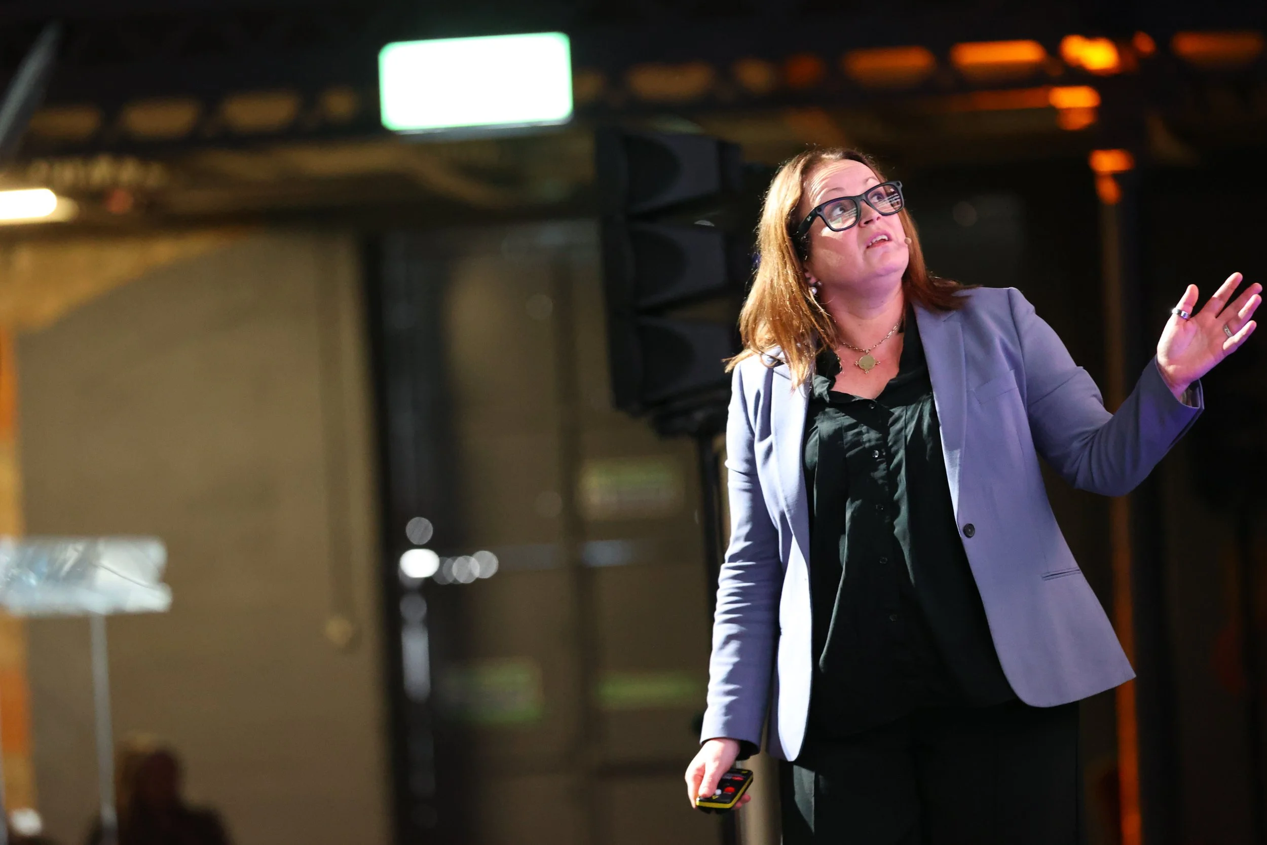 A woman with glasses, wearing a light purple blazer and black shirt, stands indoors with stage lights in the background. She is holding a remote and gesturing with her right hand.