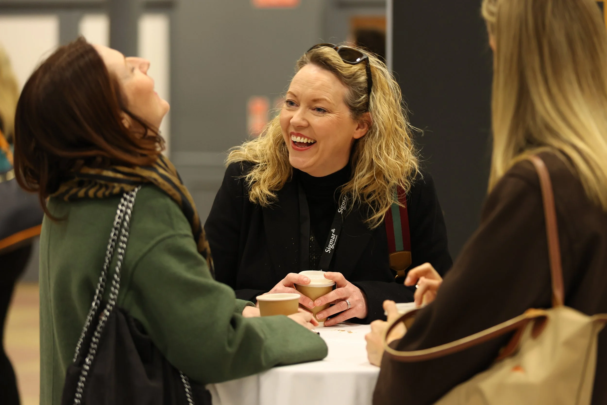 Three women smiling and talking at a round table with coffee cups at a social or networking event.