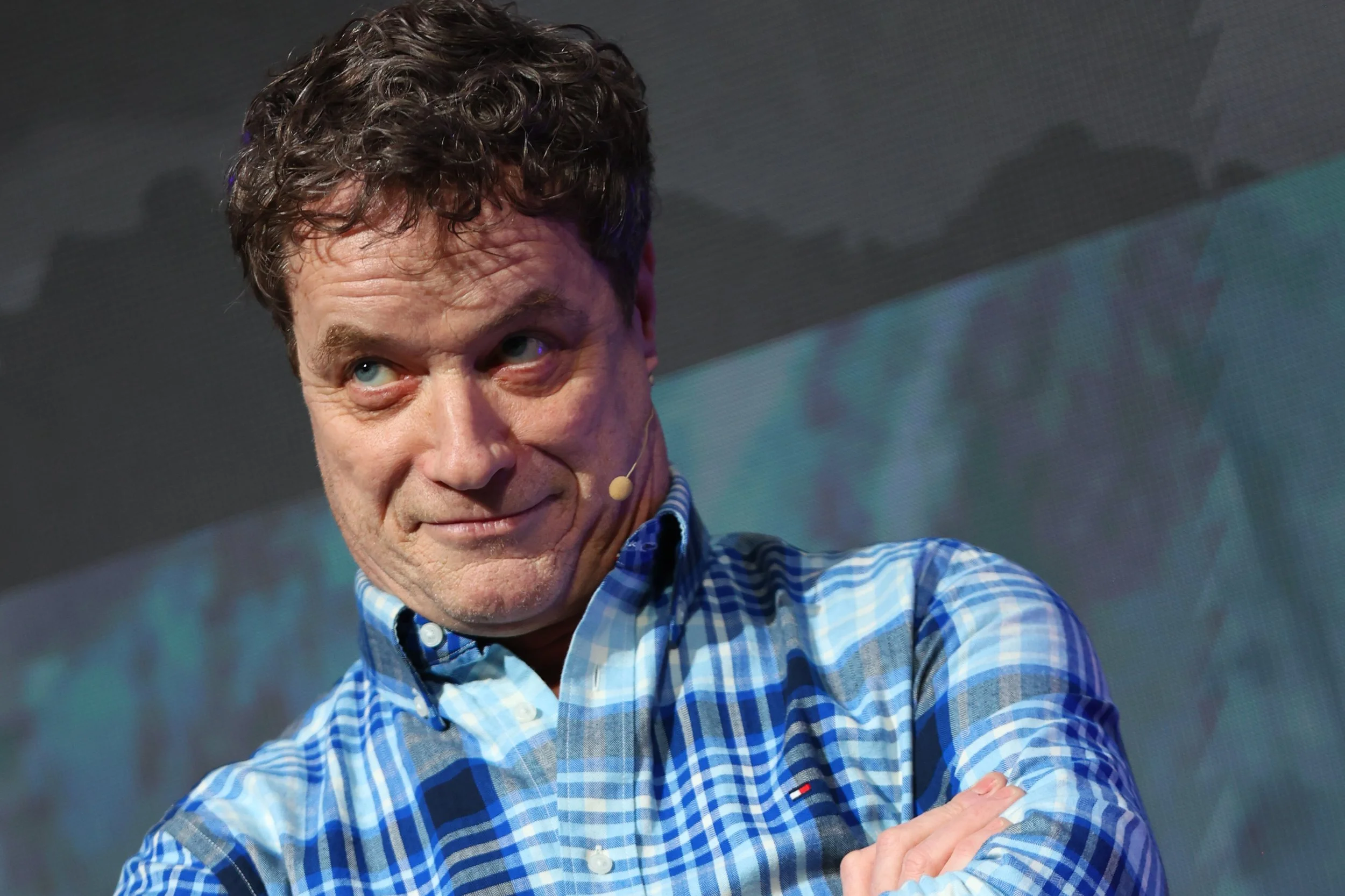 A man with curly dark hair wearing a blue and white checkered shirt, crossing his arms and looking to the side with a playful expression.
