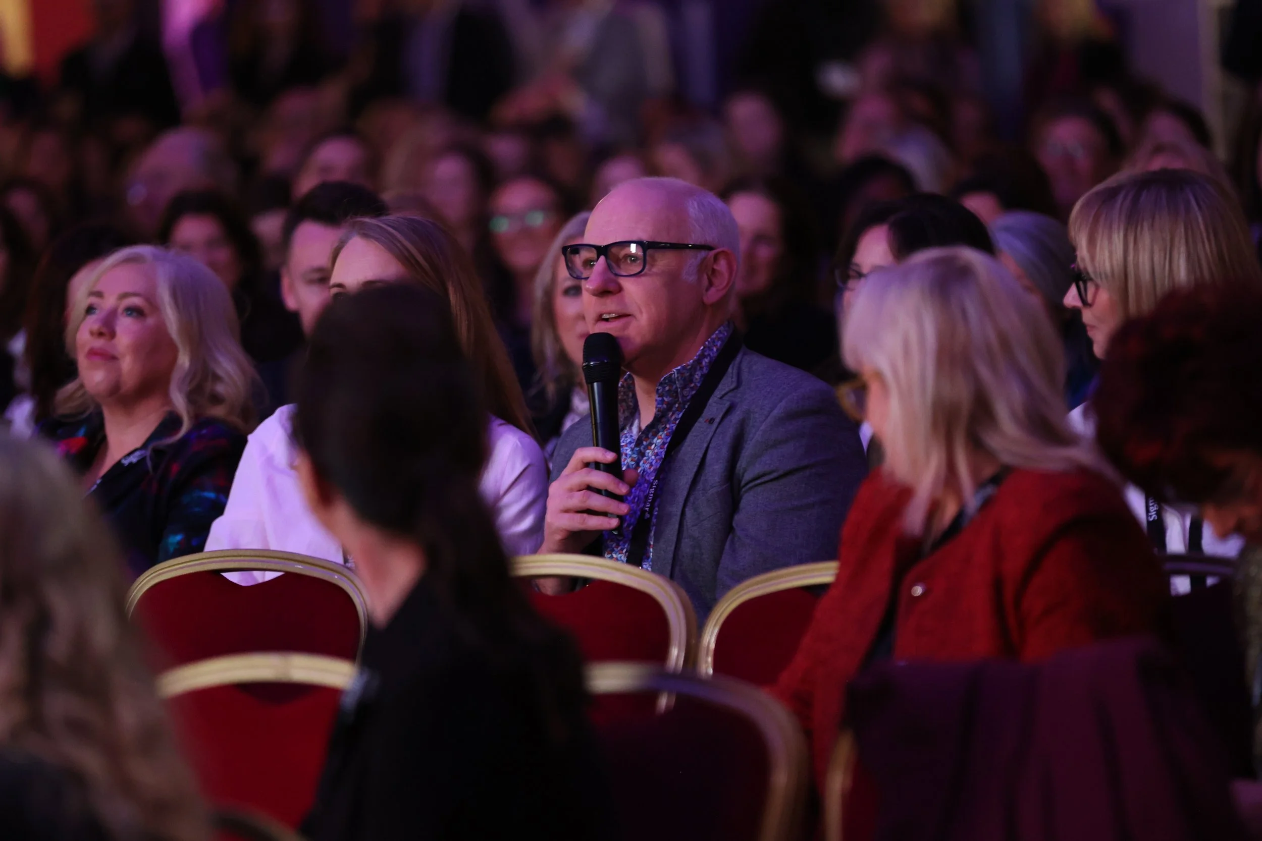 An older man with glasses holding a microphone during a conference or event, surrounded by seated attendees in a dimly lit venue.