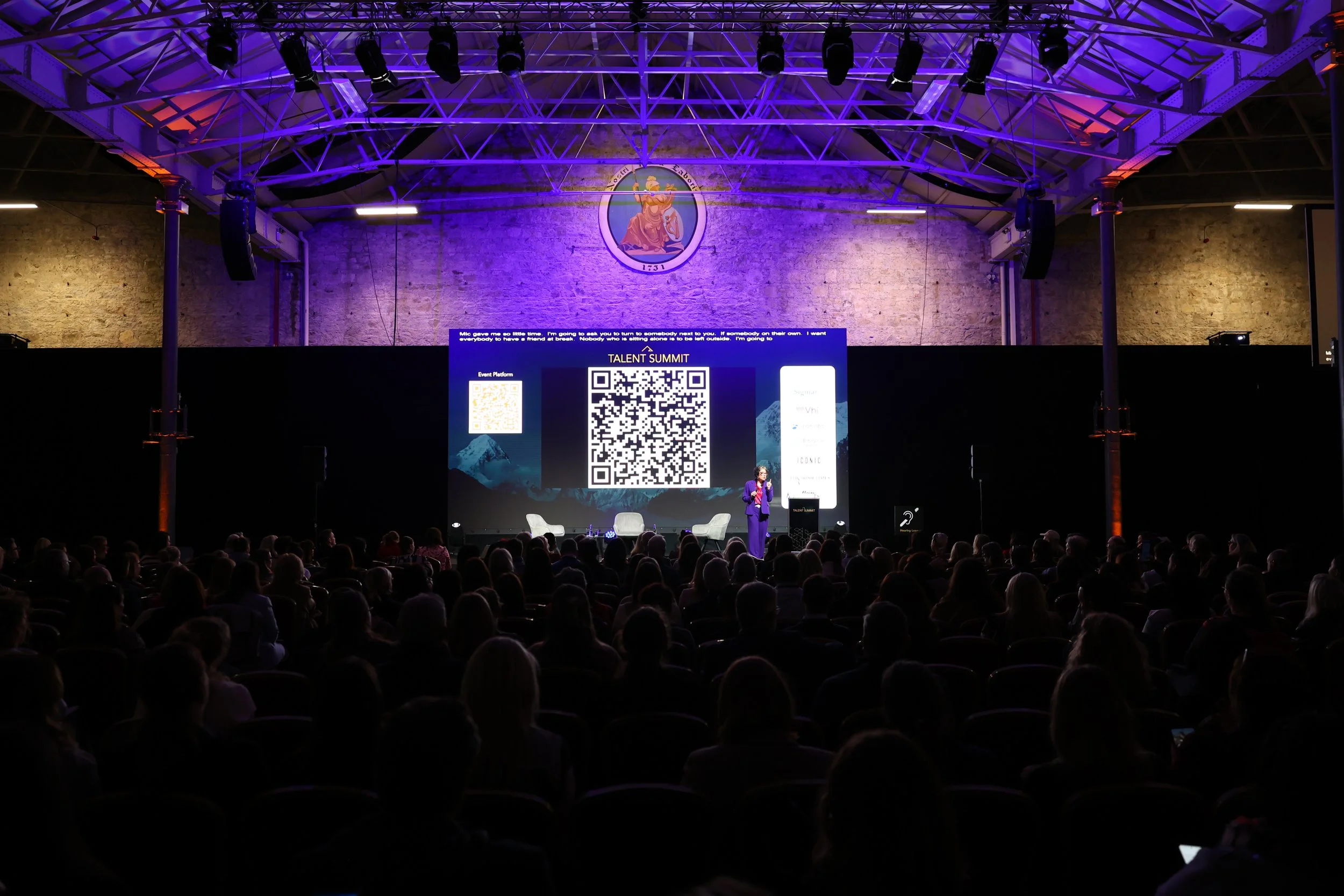 A woman in a purple suit speaking on stage at a conference with a large screen displaying a QR code and event details behind her. The audience is seated in front, and the venue has a high ceiling with lighting and brick walls.