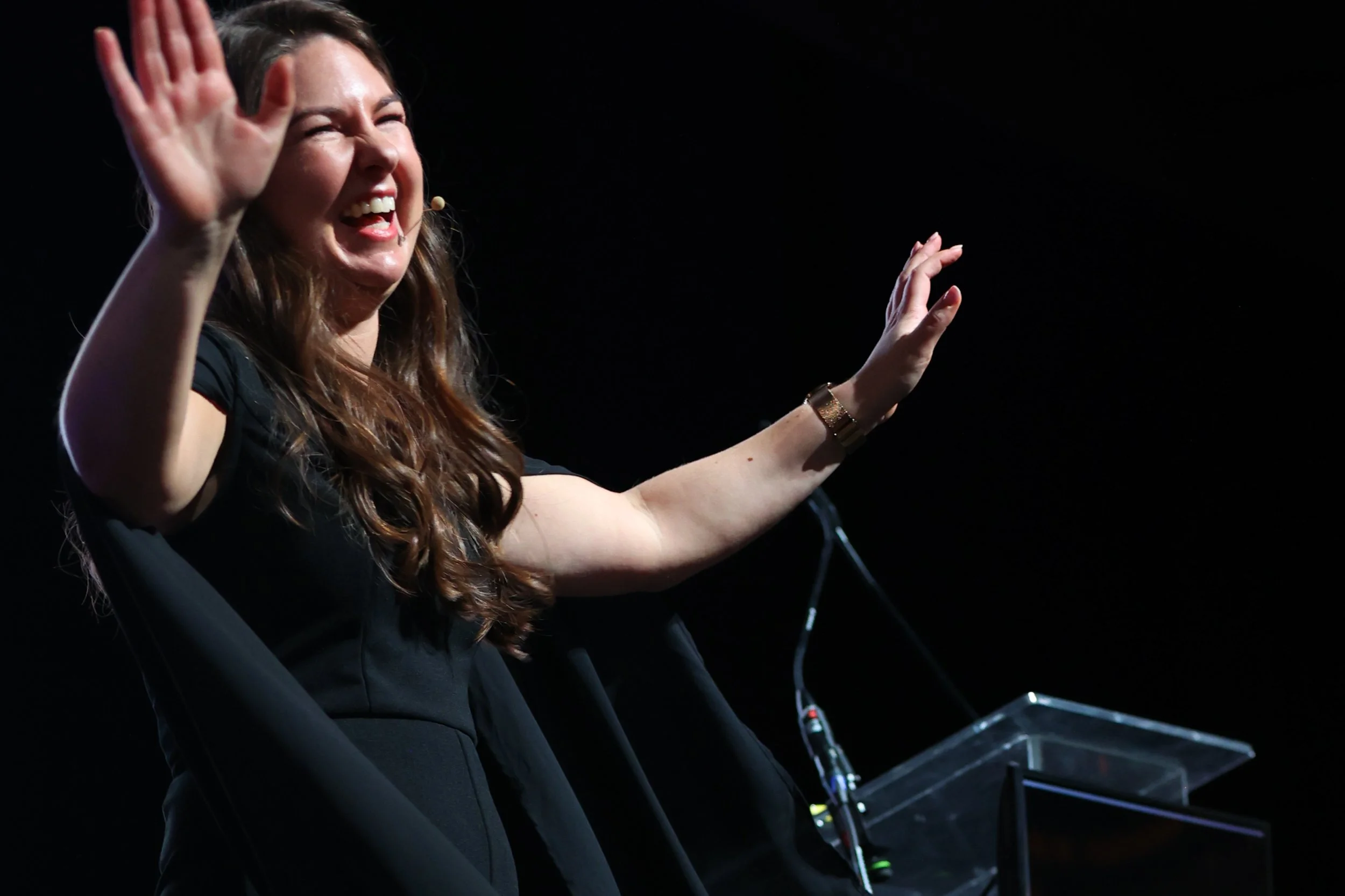 A woman with long brown hair smiling and waving while standing at a podium on a dark stage.