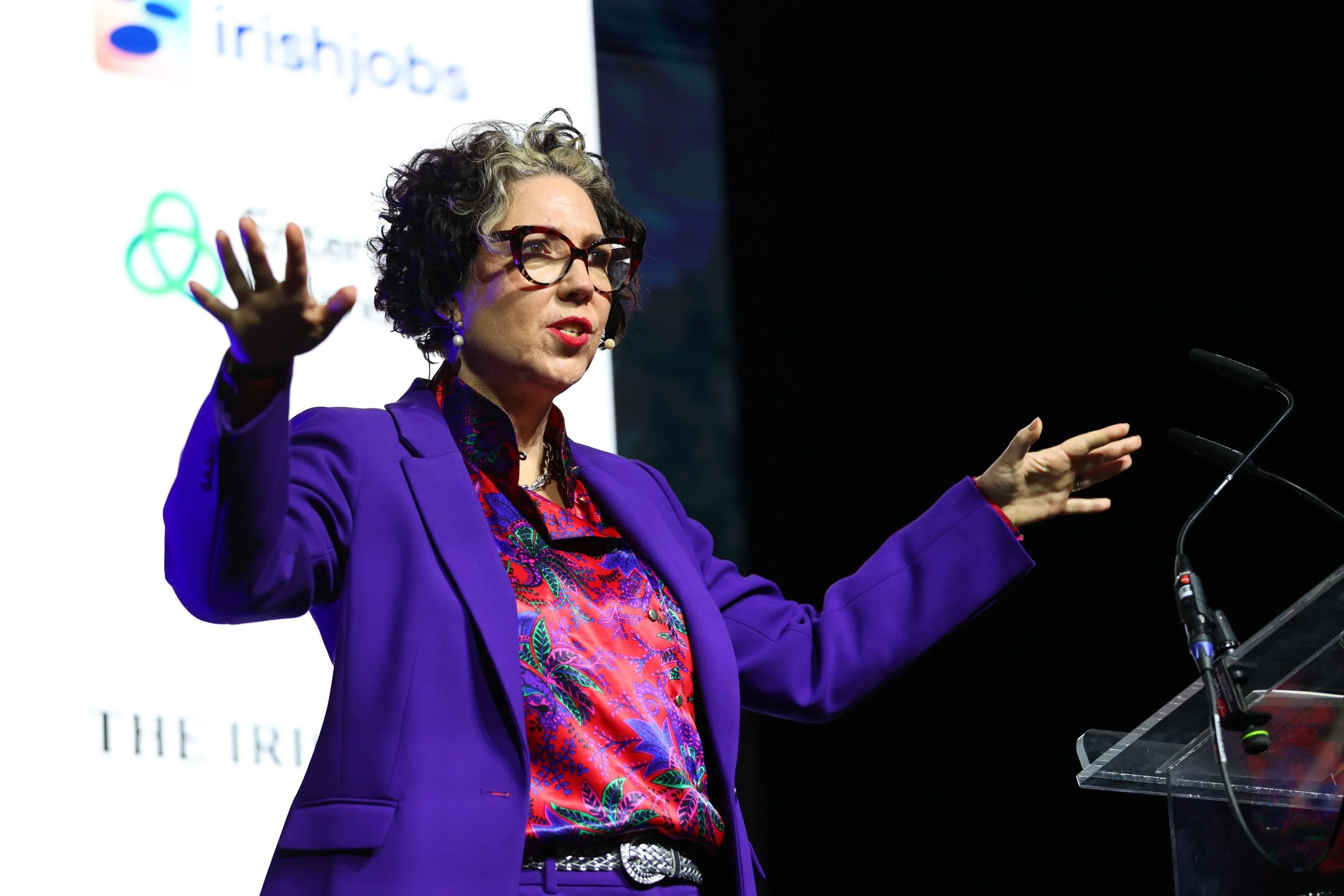 A woman with curly hair and glasses speaking at a podium during a presentation, with a screen displaying a logo behind her.