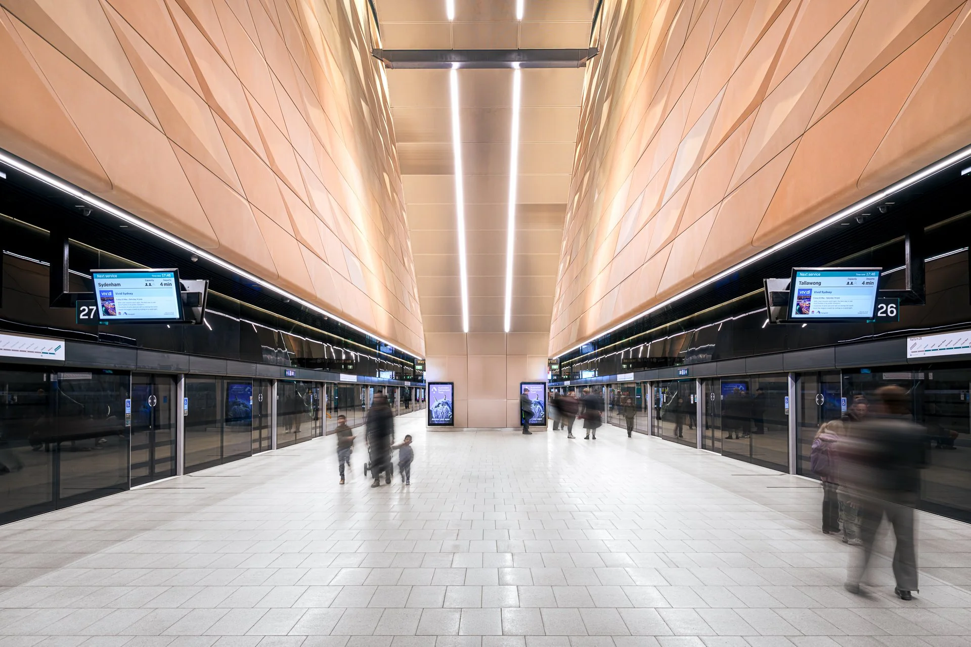 Central Metro Station with two digital departure boards, glass doors, and people walking inside, reflecting movement. Photo by Henry Li Photography.