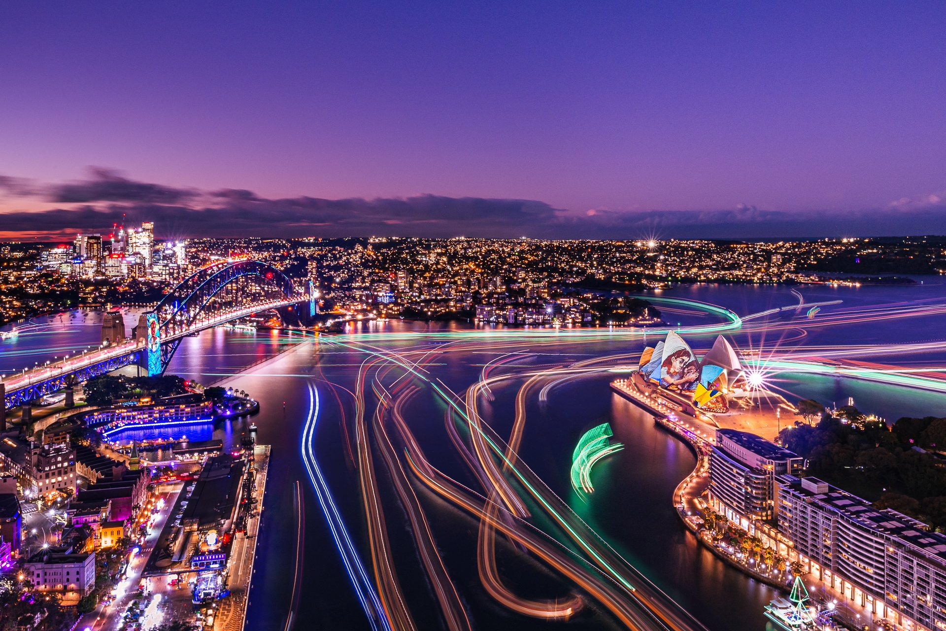 Long exposure night view of Sydney, Australia, showing the Sydney Harbour Bridge, Sydney Opera House, and city skyline with light trails from boats and traffic.