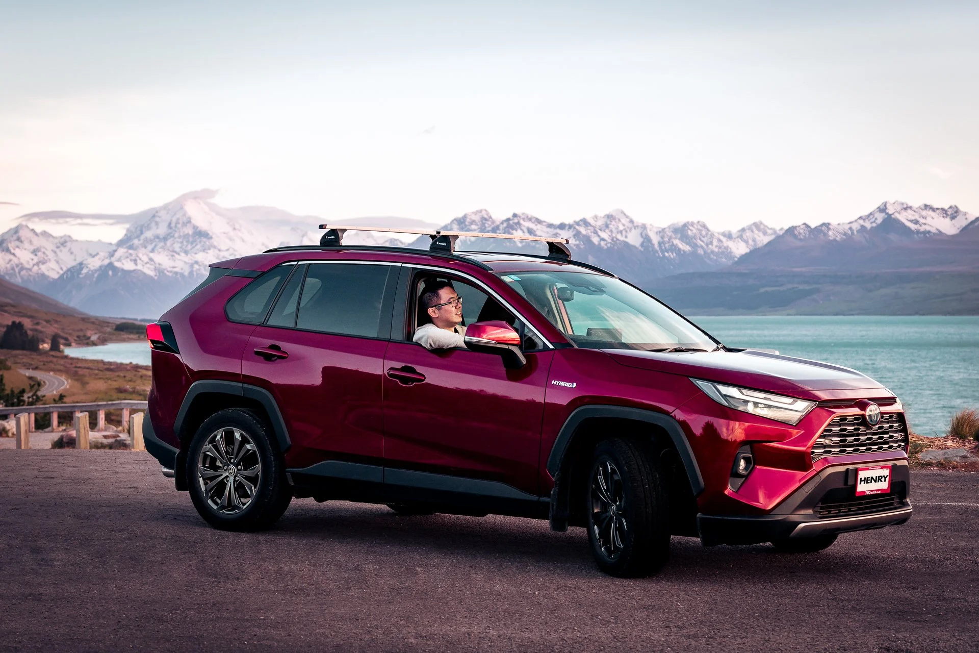 A red Toyota RAV4 hybrid SUV parked near a lake with snow-capped mountains in the background, a man is sitting in the driver's seat looking out.