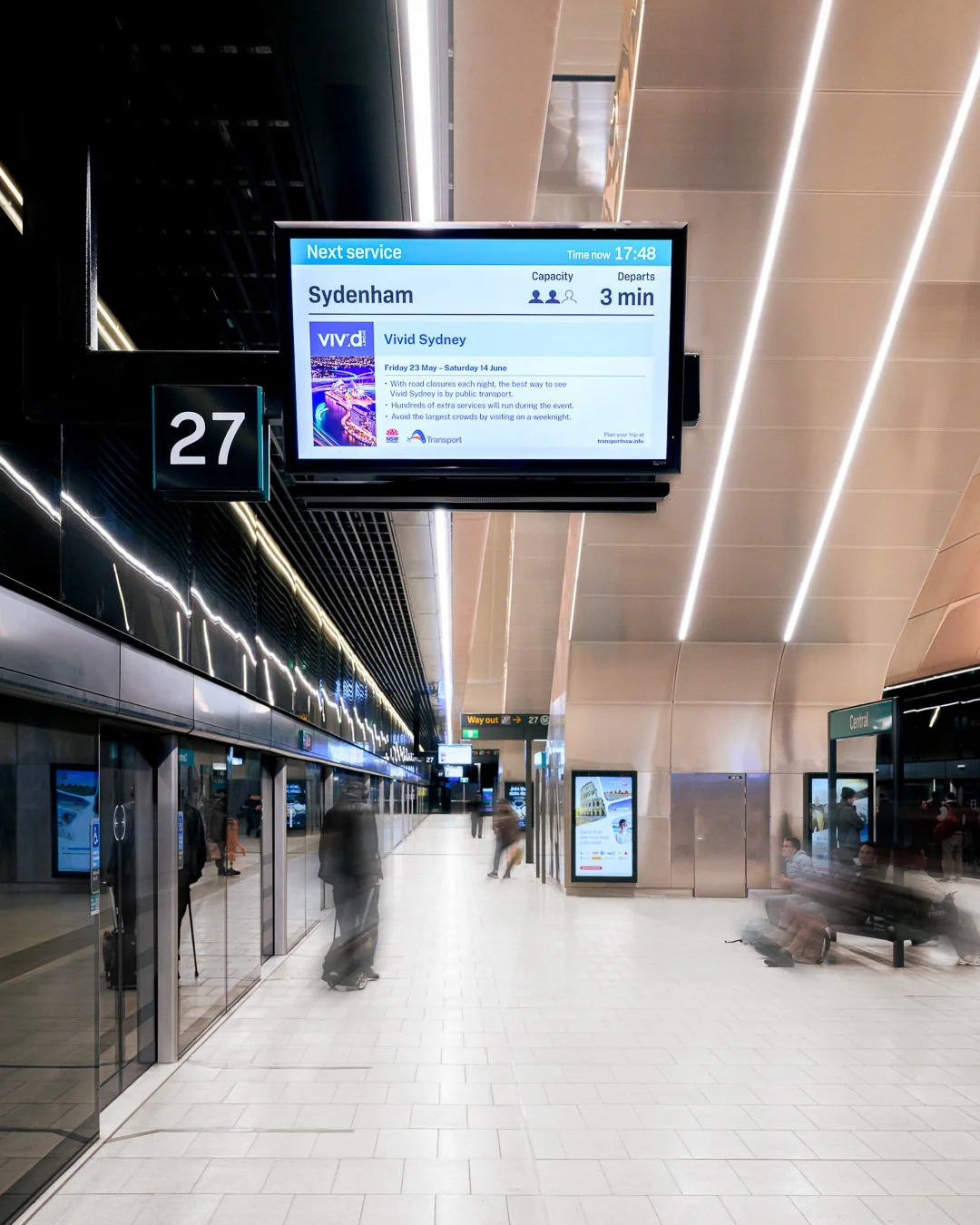 Sydney train station platform with digital timetable displaying next train to Sydenham, departure in 3 minutes, and a large screen showing train service details. Passengers are walking or sitting, with a sign indicating platform 27 and a 'Way out' si