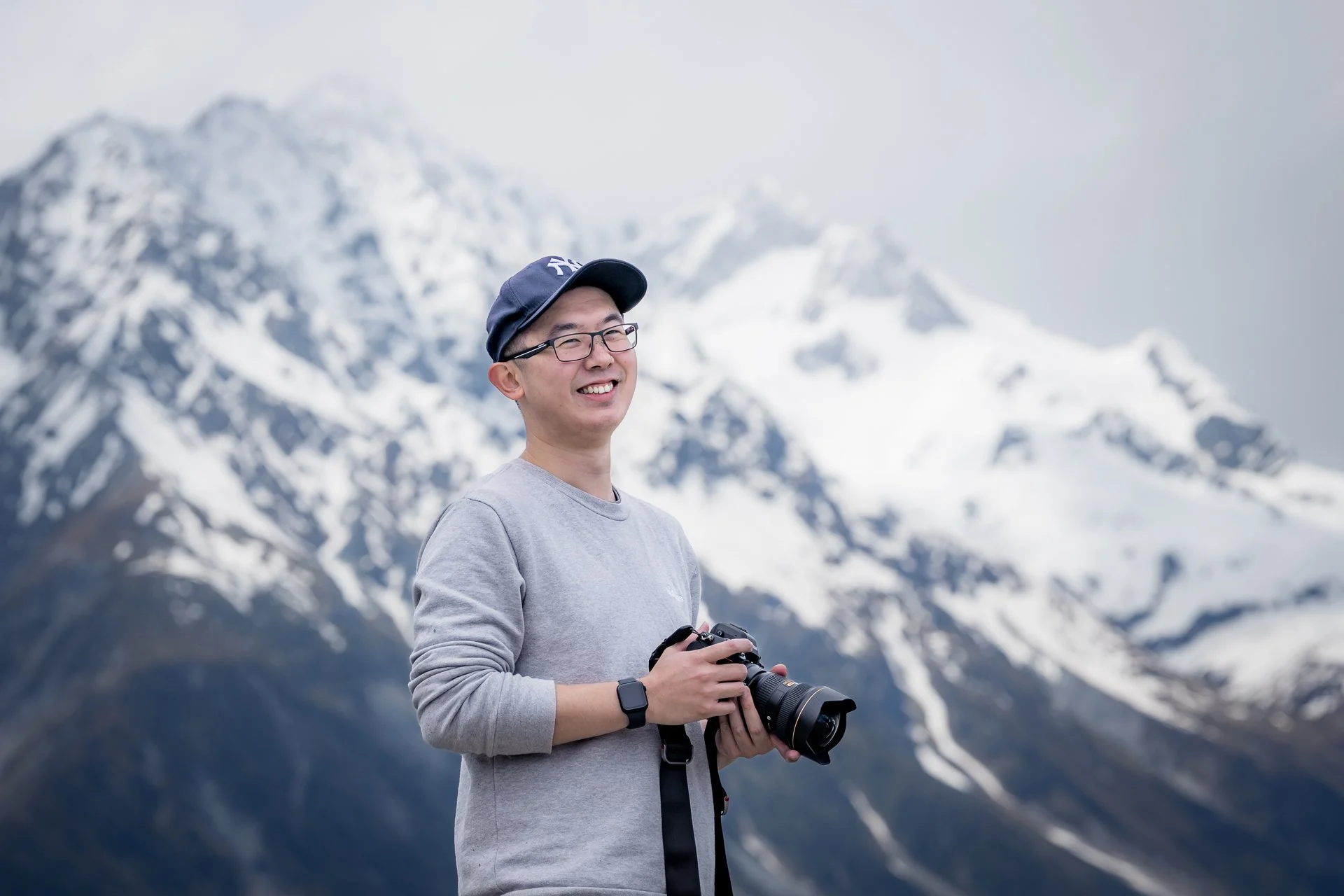 A man smiling and holding a camera with snow-capped mountains in the background.