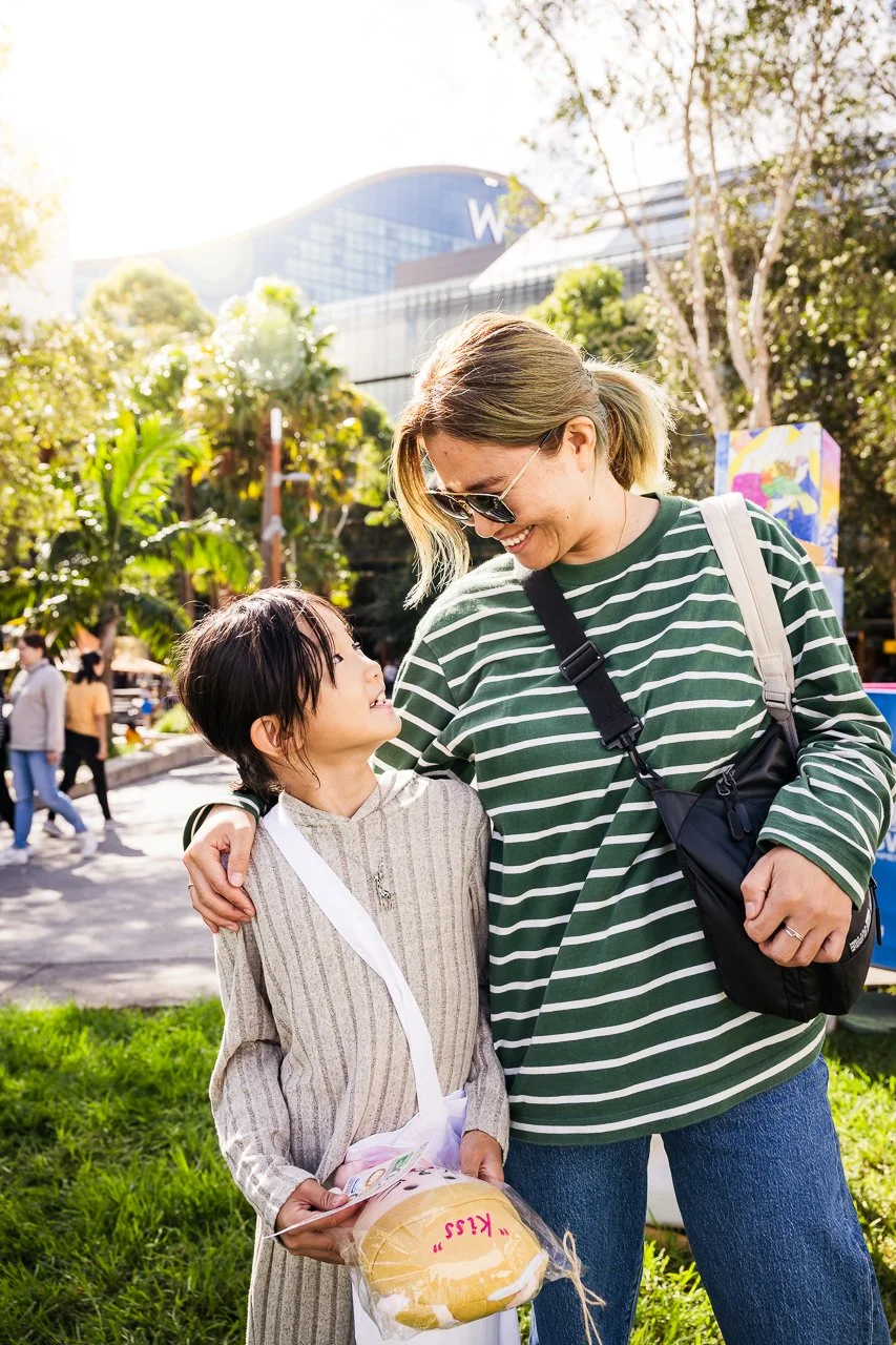 A woman and a young girl are smiling and looking at each other outdoors. The girl holds a packaged bread roll labeled 'Kiss' and some money. The woman has her arm around the girl's shoulders, and they are standing on a grassy area with trees and a modern building in the background.