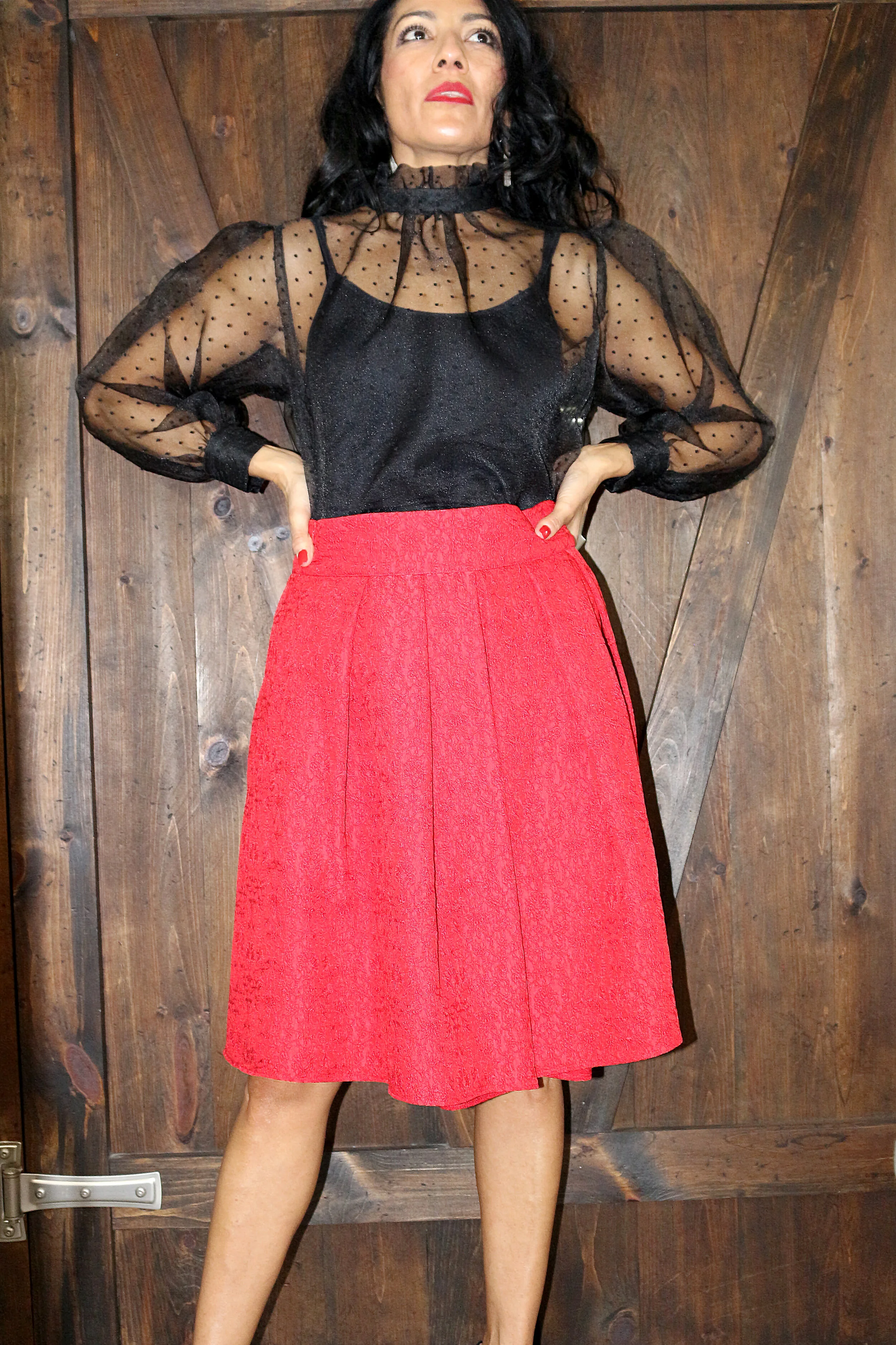 Woman in black sheer top and red skirt standing against a wooden wall.