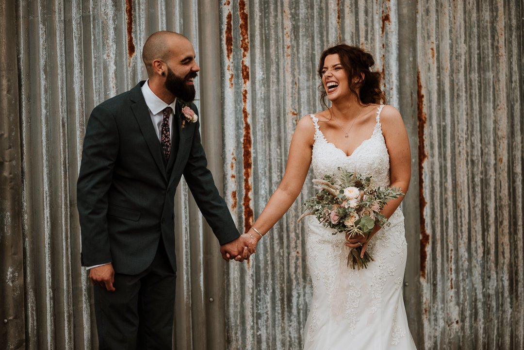 groom and bride are stood in front of a corrugated iron fence, both are in fits of laughter seemingly at a joke that was just told