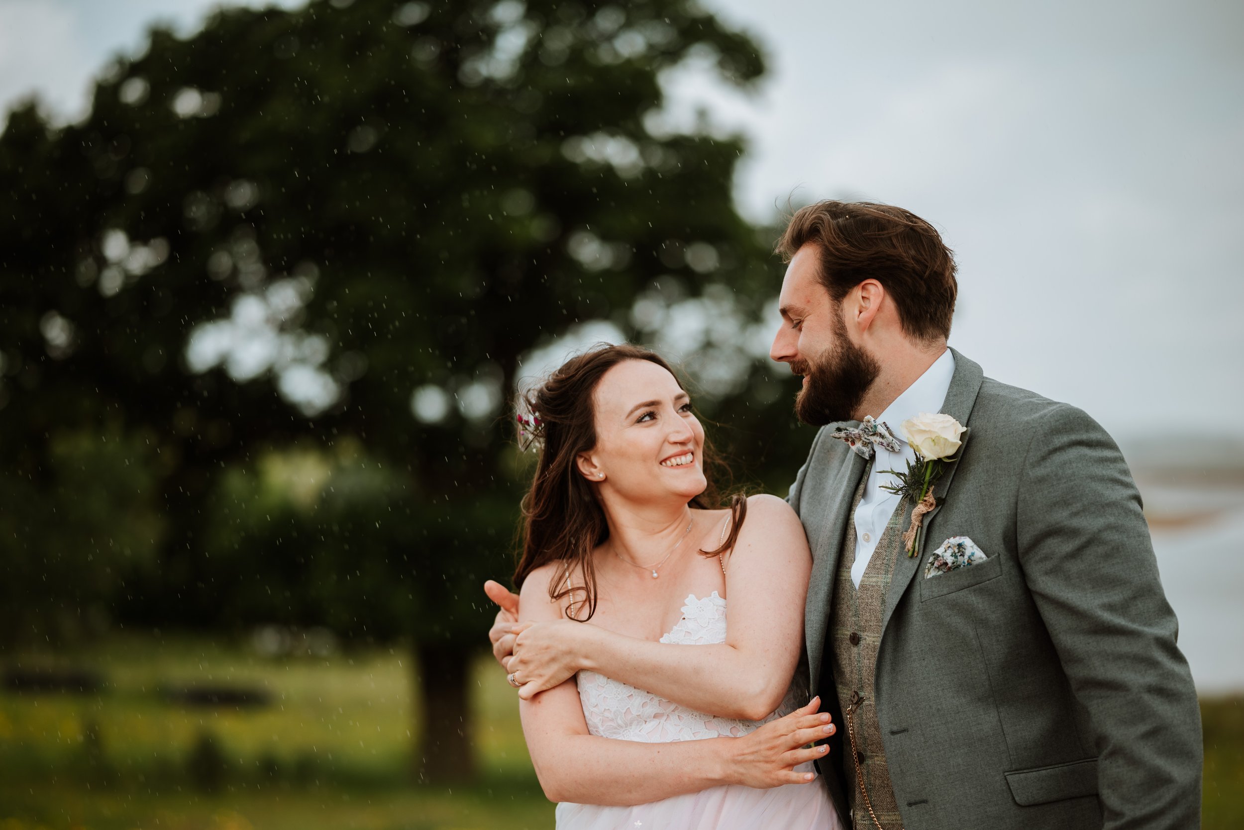 a groom has his arm around his bride as they smile at each other despite it being rainy outside