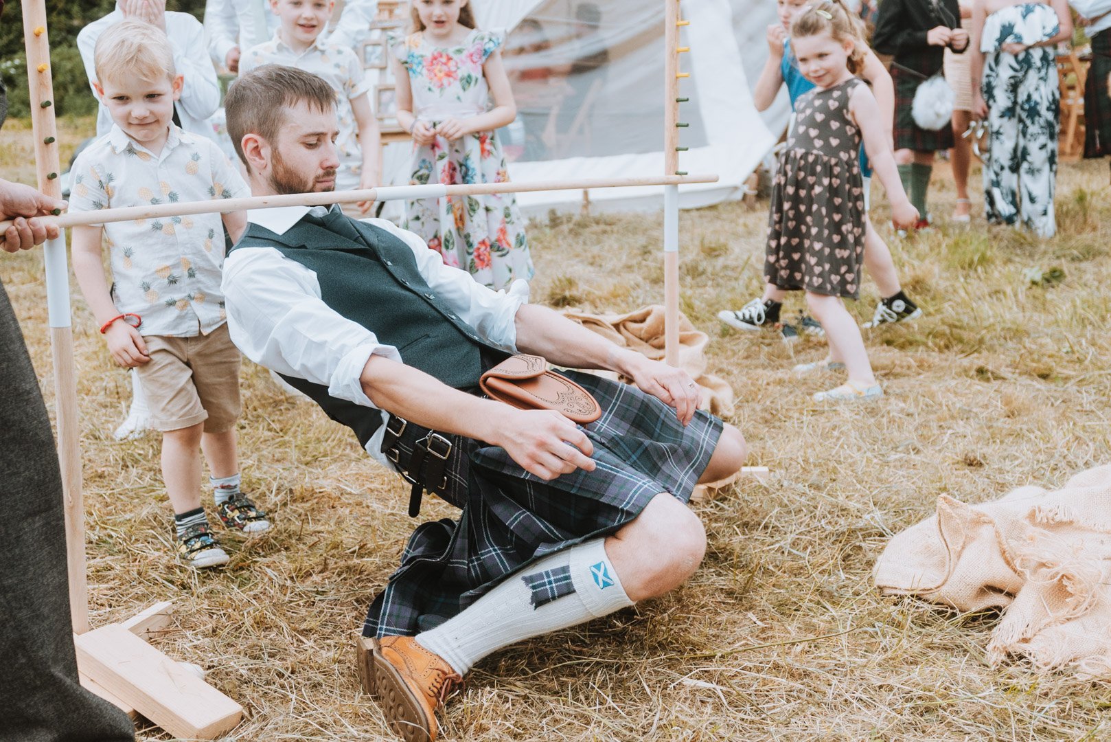 A groom dressed in a kilt and smart shirt attempts the limbo on a camping field in Somerset