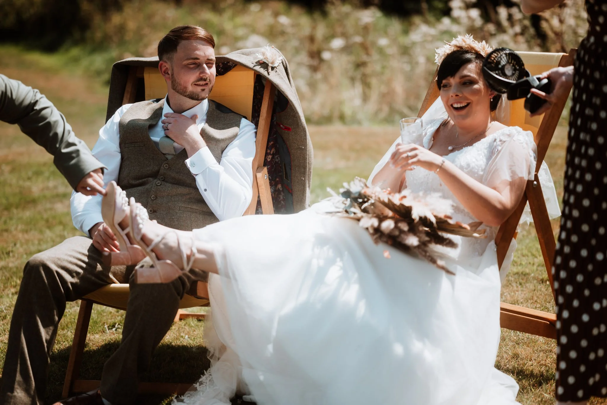 bride and groom are sat on lawn furniture, she has her feet up, is laughing and someone is holding a fan next to her face to cool her down. groom is looking over to the bride