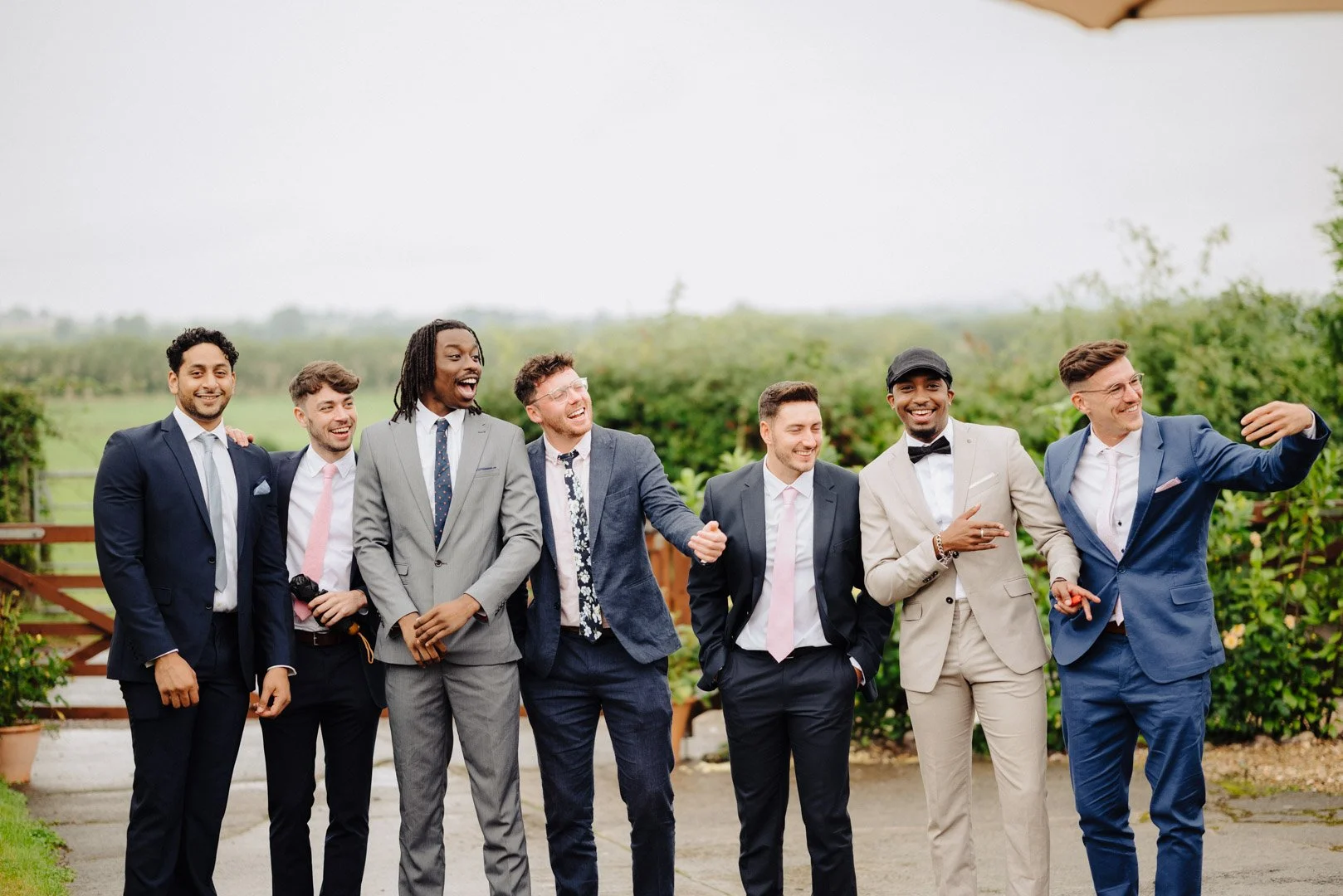 Groomsmen and other male wedding guests stand in a line for a candid group photo and some of the men are waving the groom over to them out of picture. at Dairyhouse Farm Wedding Barn Venue, Somerset