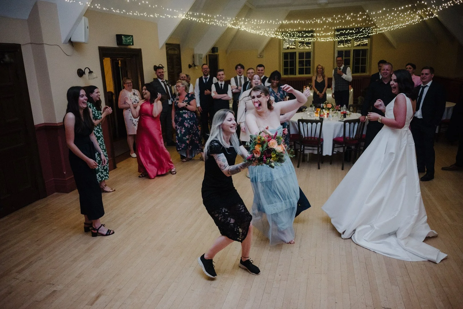 wedding guest catches the flowers on the dance floor while other guests stand around watching. The bride and other guests are laughing at the catch