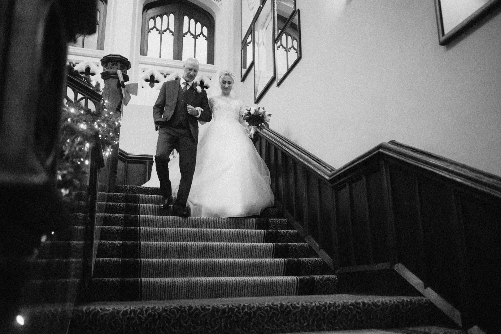 black and white image of a bride being escorted down a grand stair case at De Vere Tortworth Court, on her way to the ceremony
