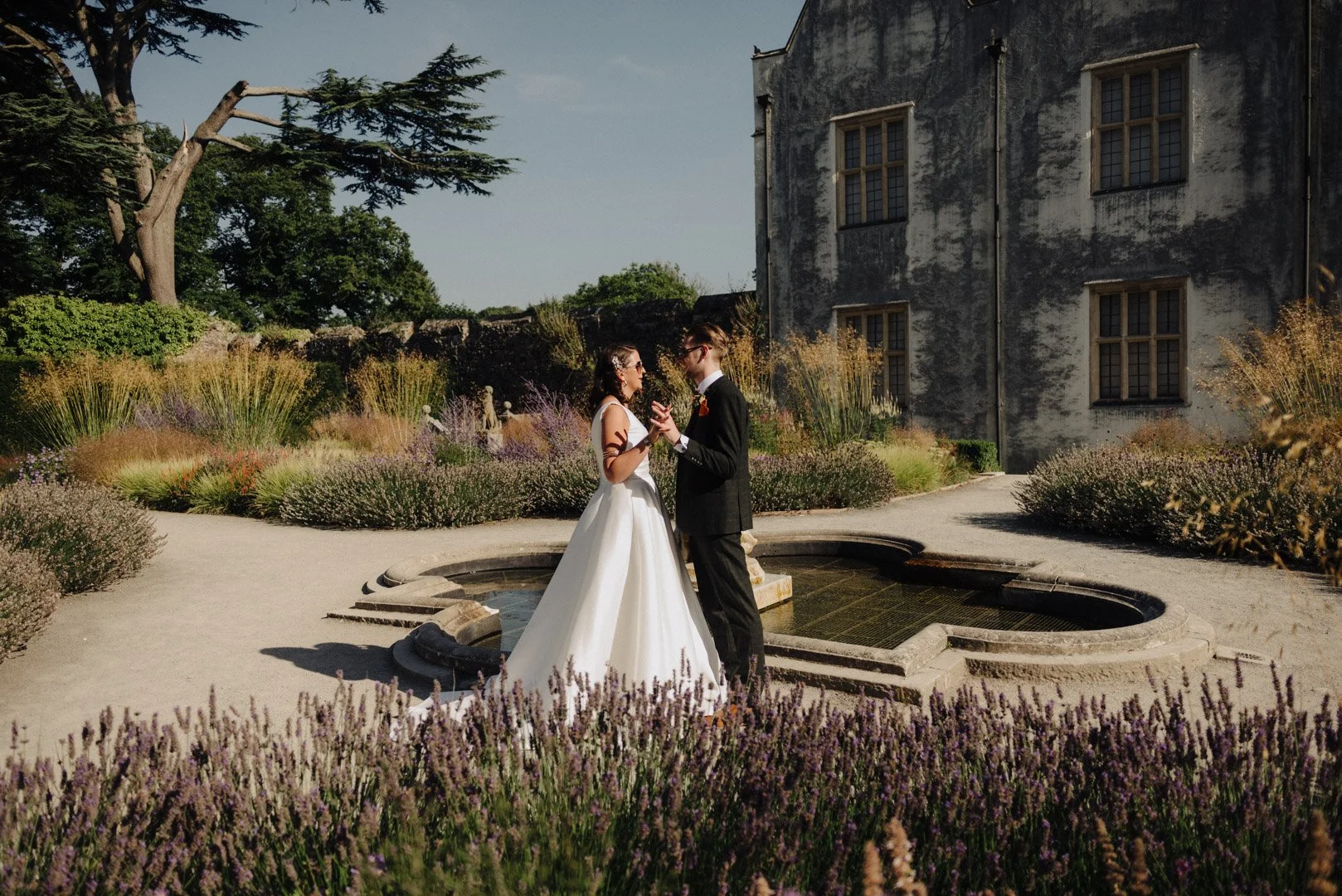 a bride and groom hold hands and take a moment to speak and relax during their wedding, they are stood in the beaitiful gardens of St Fagans in Wales during a really sunny day. they are wearing sunglasses.