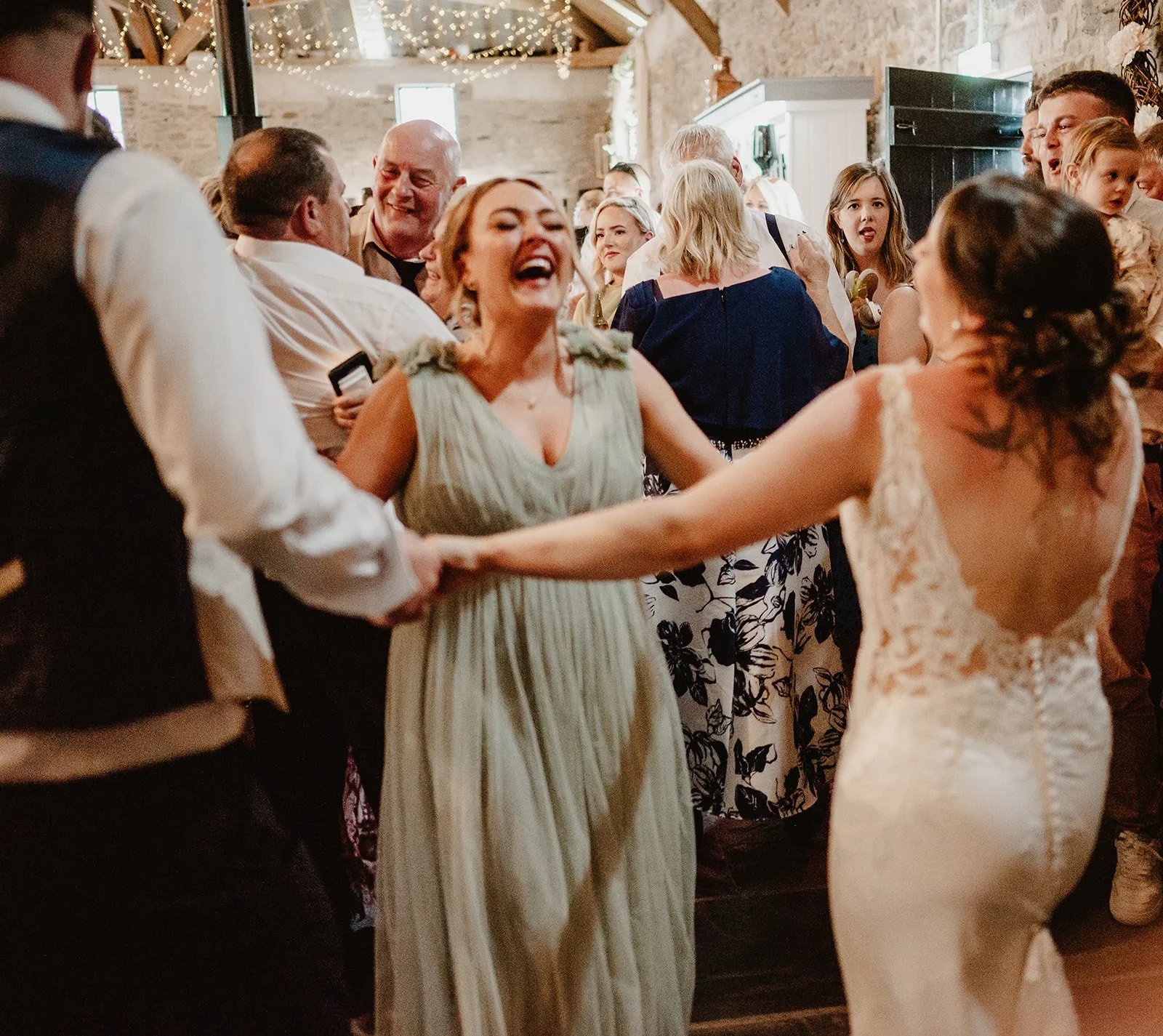 a really busy and packed dance floor at a wedding with lots of people smiling and dancing, including the bride holding hands with one of the bridesmaids as they laugh and dance