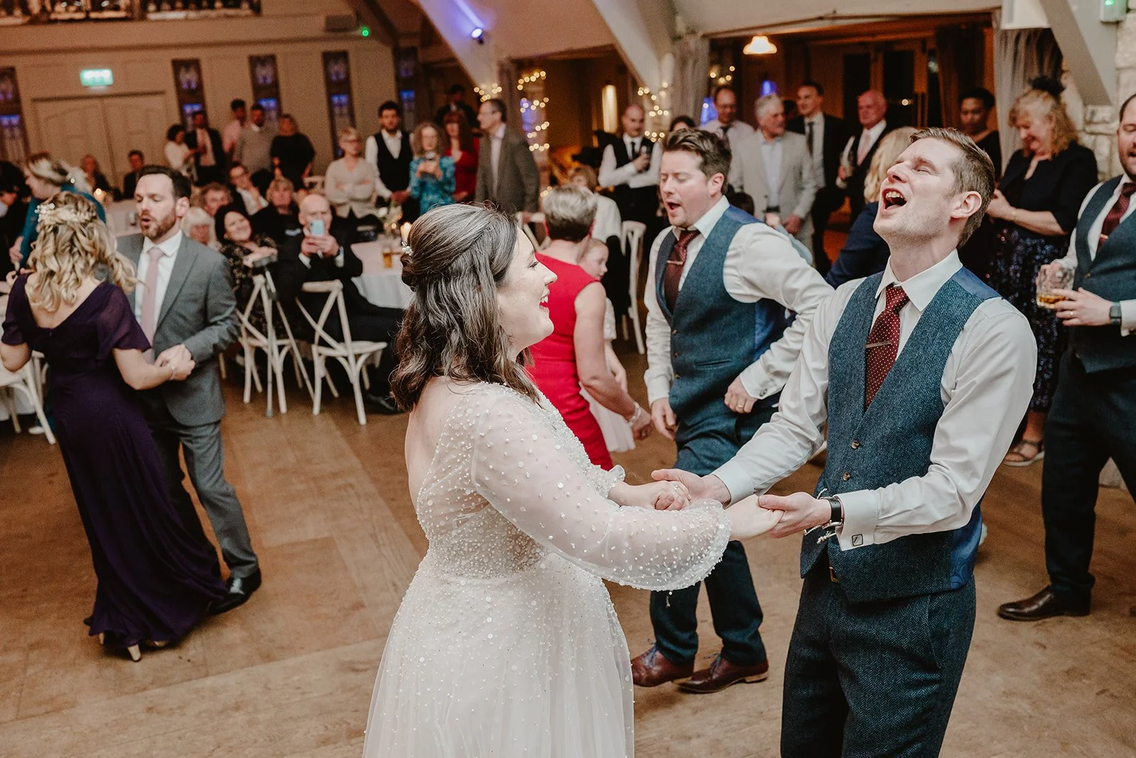 a bride and groom hold hands and dance, singing along to the words of a song that is playing during their wedding reception in Bristol and behind them their guests are also dancing