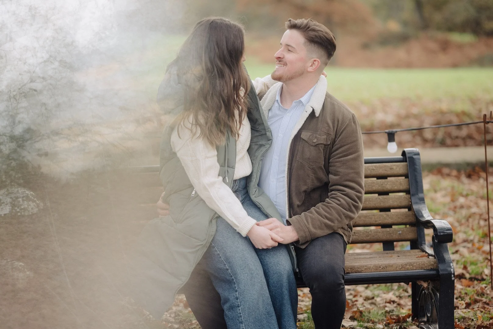Engaged couple sit on park bench, woman on the man's lap and they are looking at each other smiling. At Margam Park In Port Talbot