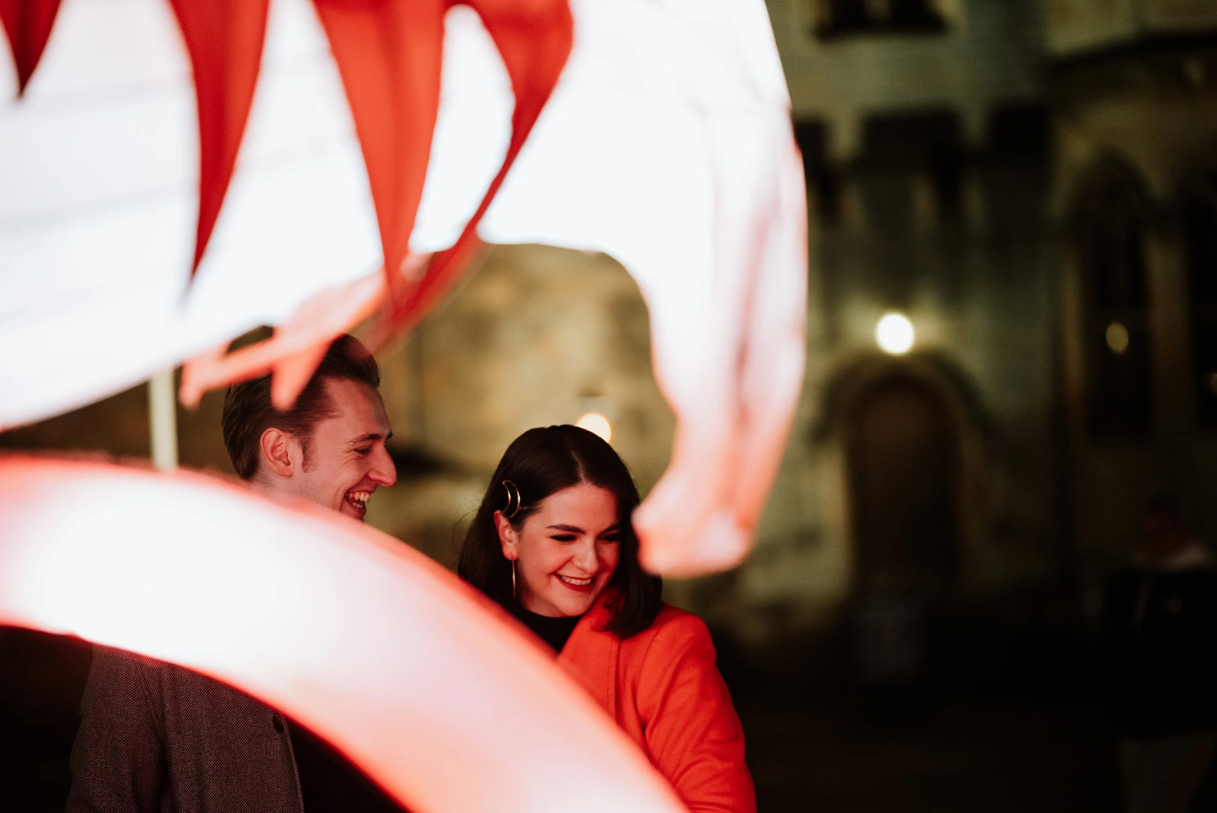 man and woman stand behind a lit up christmas themed display outside of Cardiff Castle, man is smiling and looking at the woman and she's looking away smiling bashfully, during an engagement photo shoot