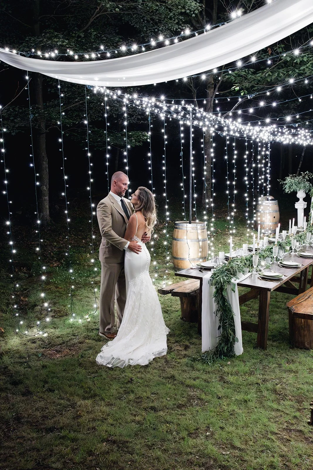 Bride and groom embrace under twinkling lights in a reception space beside a dinner table.