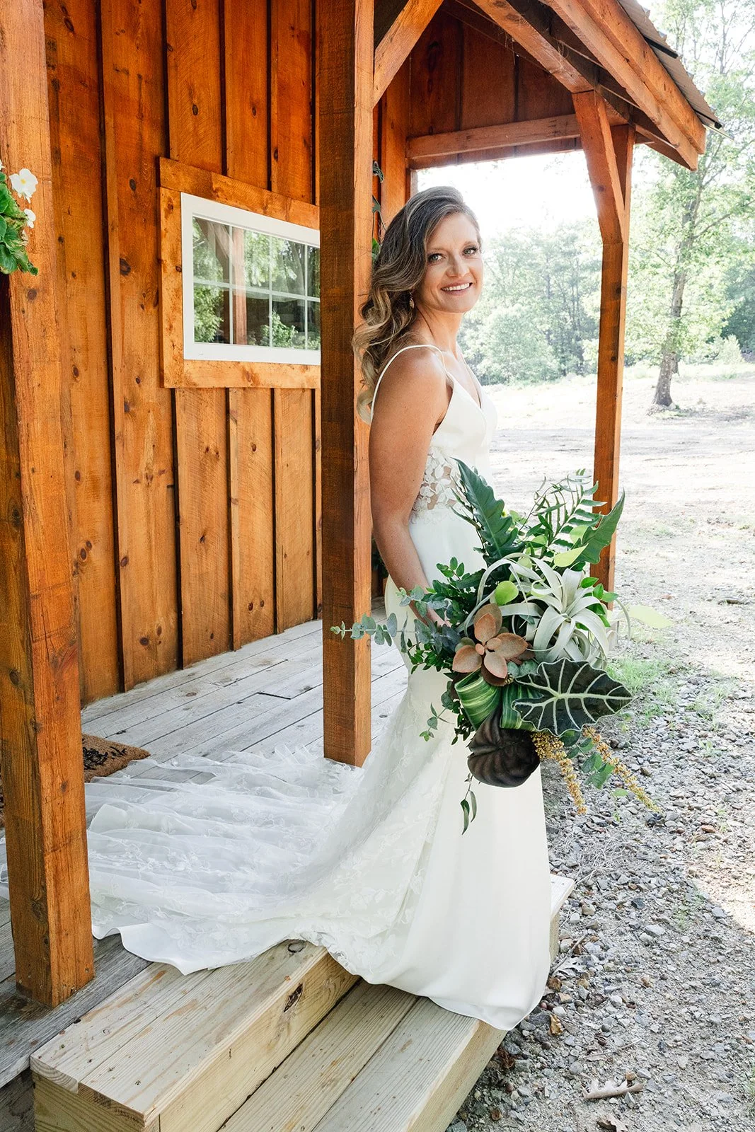 Bride emerges from private woodsy cabin in wedding dress holding greenery bouquet.