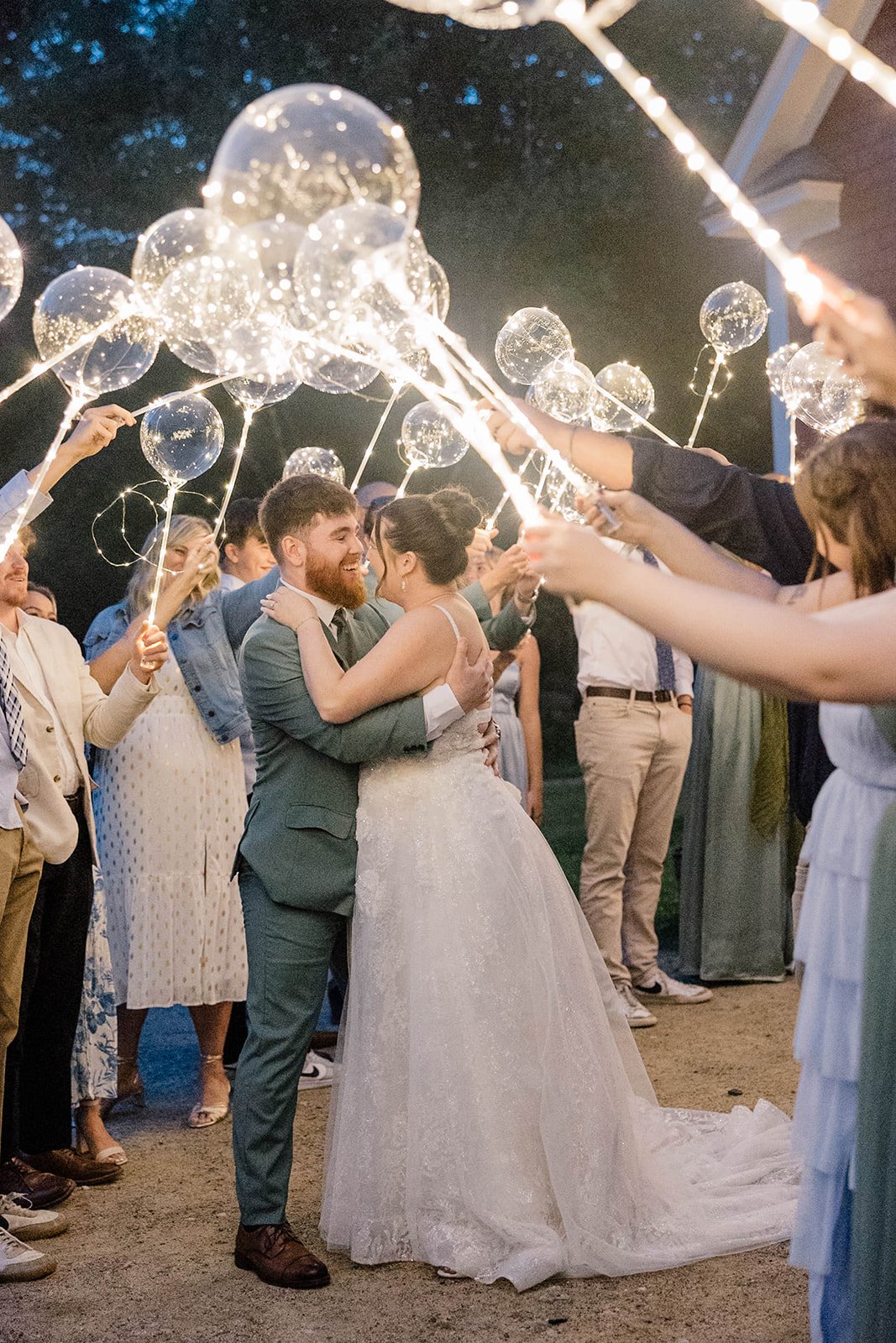 Bride and groom embrace while their loved ones create a tunnel of lighted balloons during their garden wedding send off.