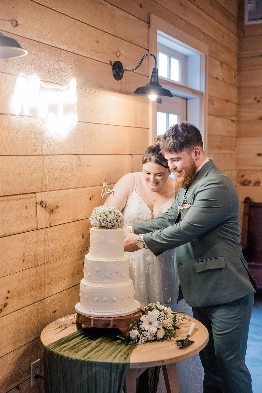 Bride and groom smile as they cut their three tiered wedding cake surrounded by flowers, carrying on the theme of their garden wedding.
