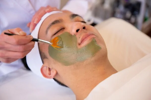 A man receiving a facial treatment with a brush applying a green mask on his face while lying down with eyes closed.