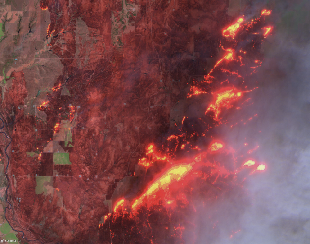 Satellite image of a large wildfire spreading, with bright red and orange areas indicating intense fire and smoke plumes.