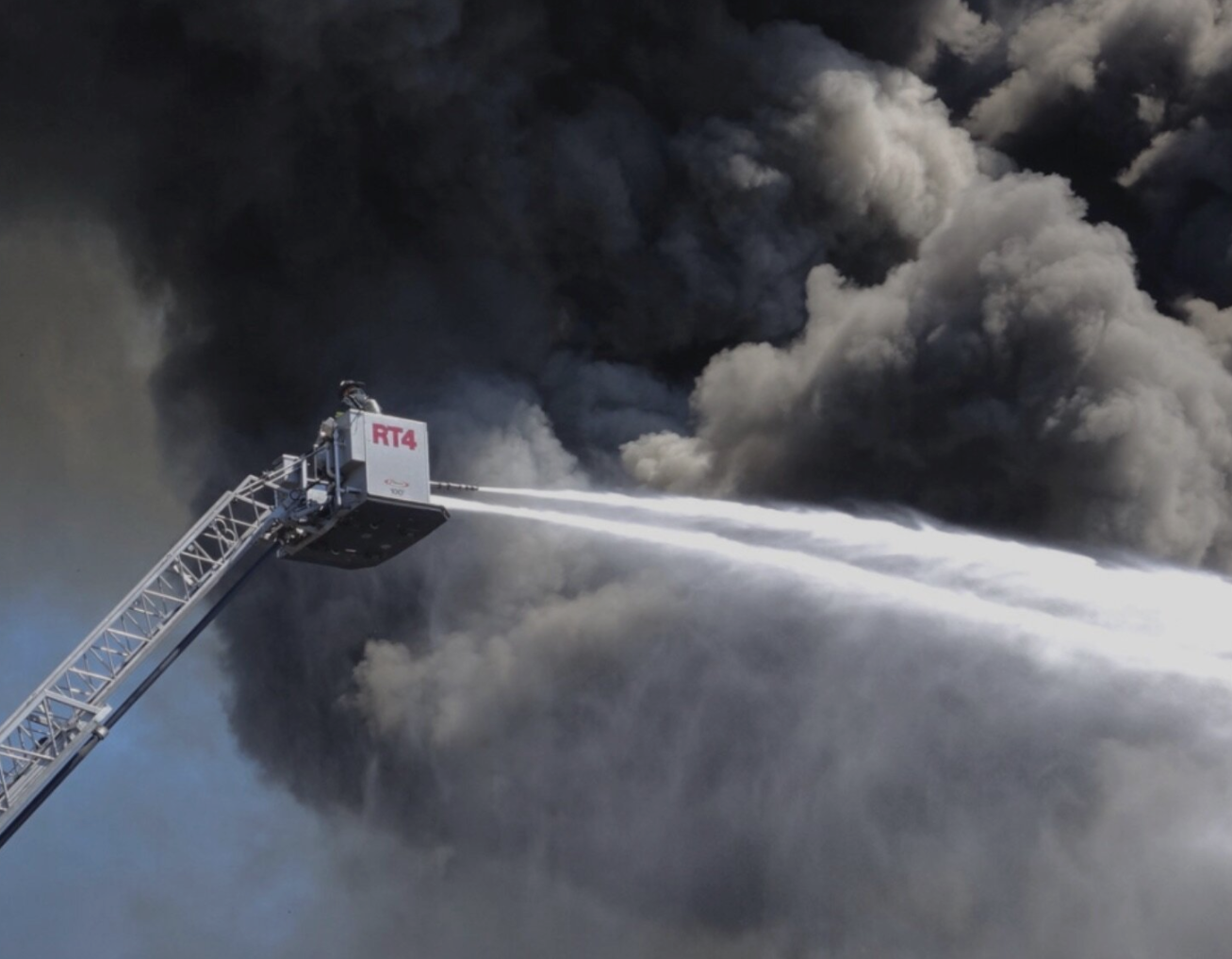 Firefighter on an elevated ladder spraying water at thick smoke clouds.
