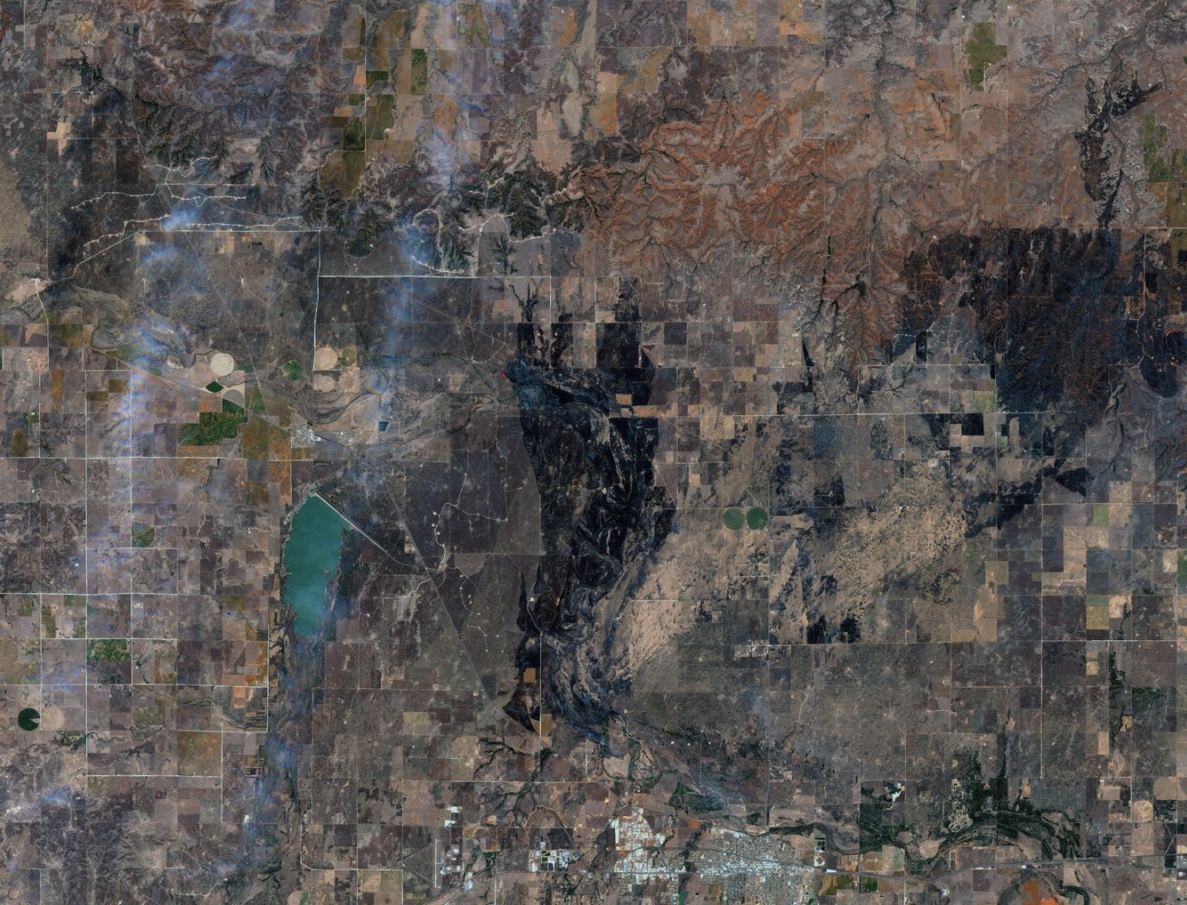 Aerial view of agricultural fields and landscape near a body of water, with a patchwork of farm plots and surrounding natural terrain. Includes visible irrigation circles and roads.