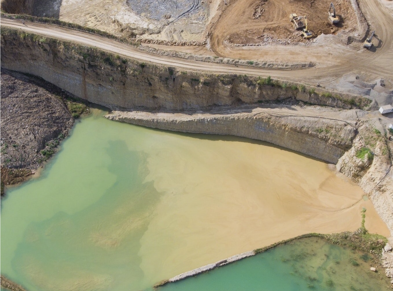 Aerial view of a mining quarry with green and brown water, surrounded by dirt roads and construction equipment.
