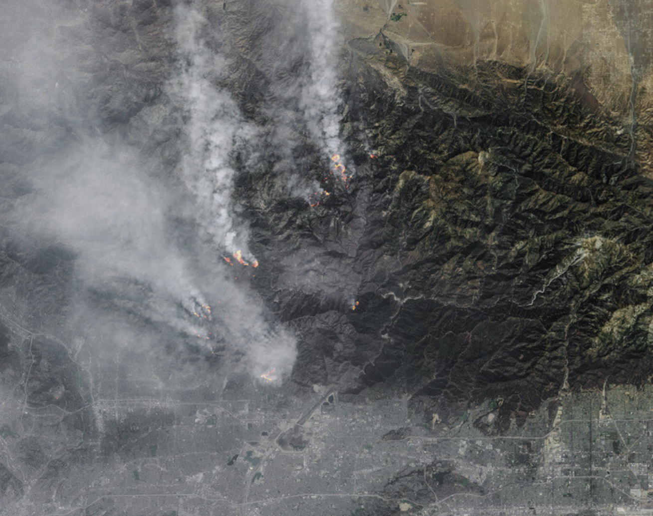 Aerial view of a wildfire in a mountainous region with smoke plumes rising over the landscape near an urban area.