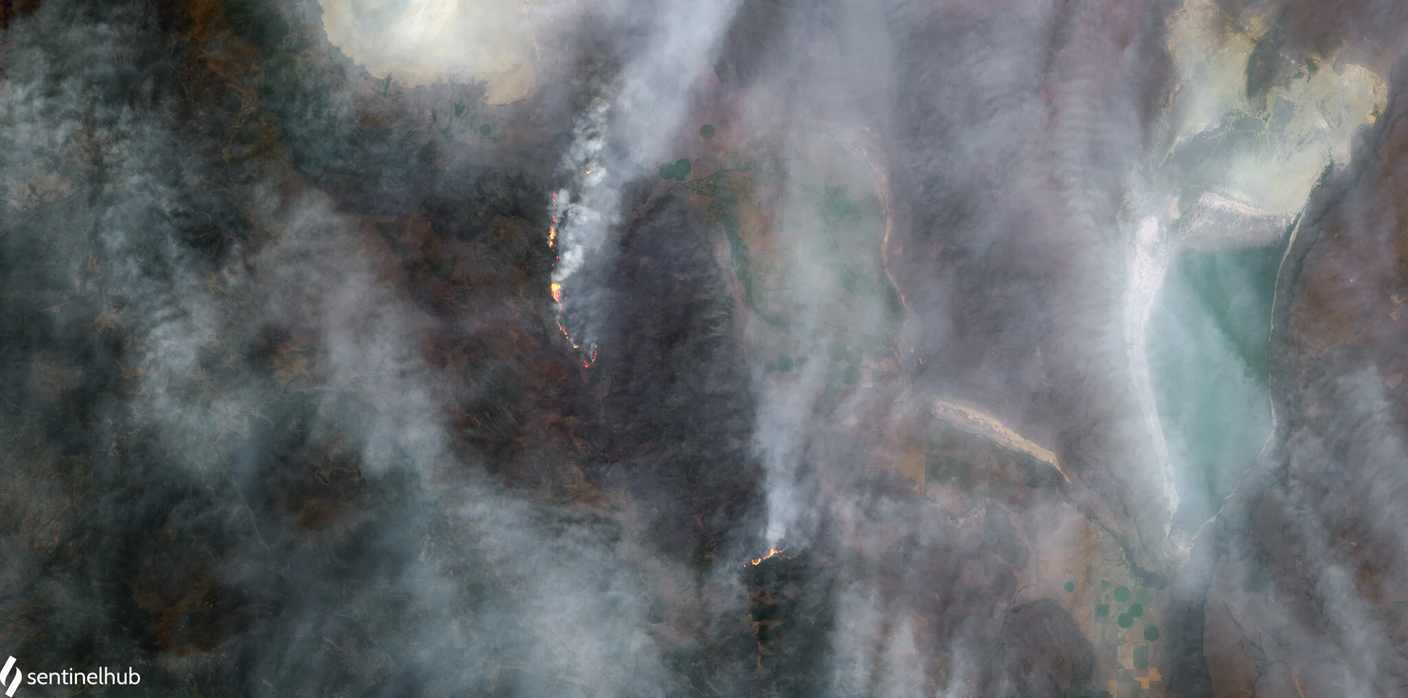 Satellite image showing a wildfire with visible smoke and flames. Surrounding areas appear to be a mix of landscapes, including green patches and a large body of water. Haze from the smoke covers parts of the image.