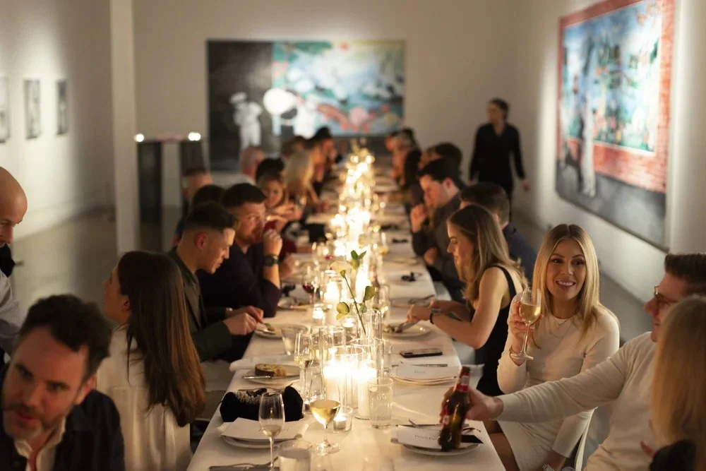 A group of people sitting at a long dinner table in an art gallery, enjoying a meal with wine and candles.